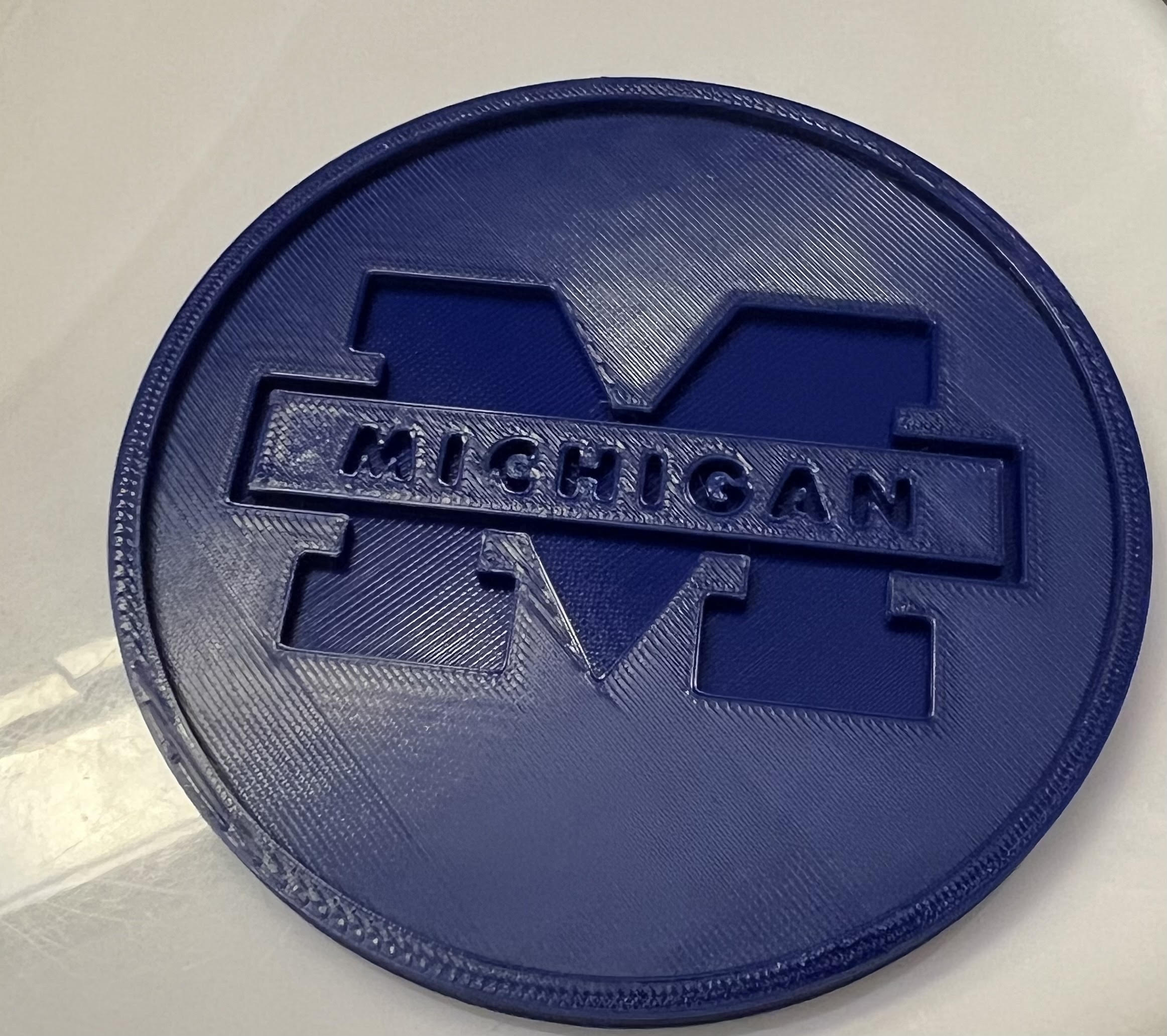 Navy blue 3D printed circular cup coaster with the Michigan logo in the center