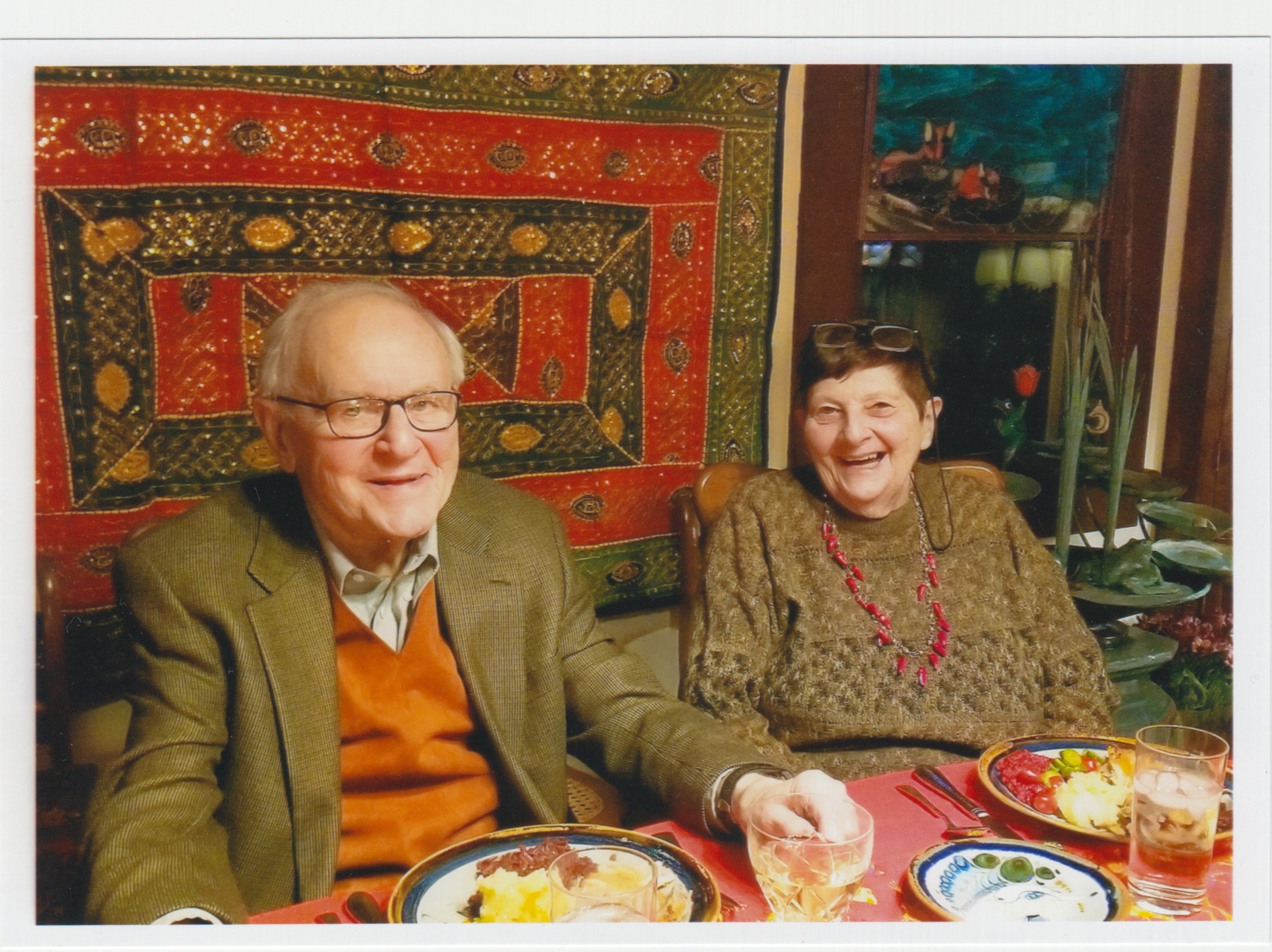 Elderly smiling couple sitting at a table, in front of a red and green wall