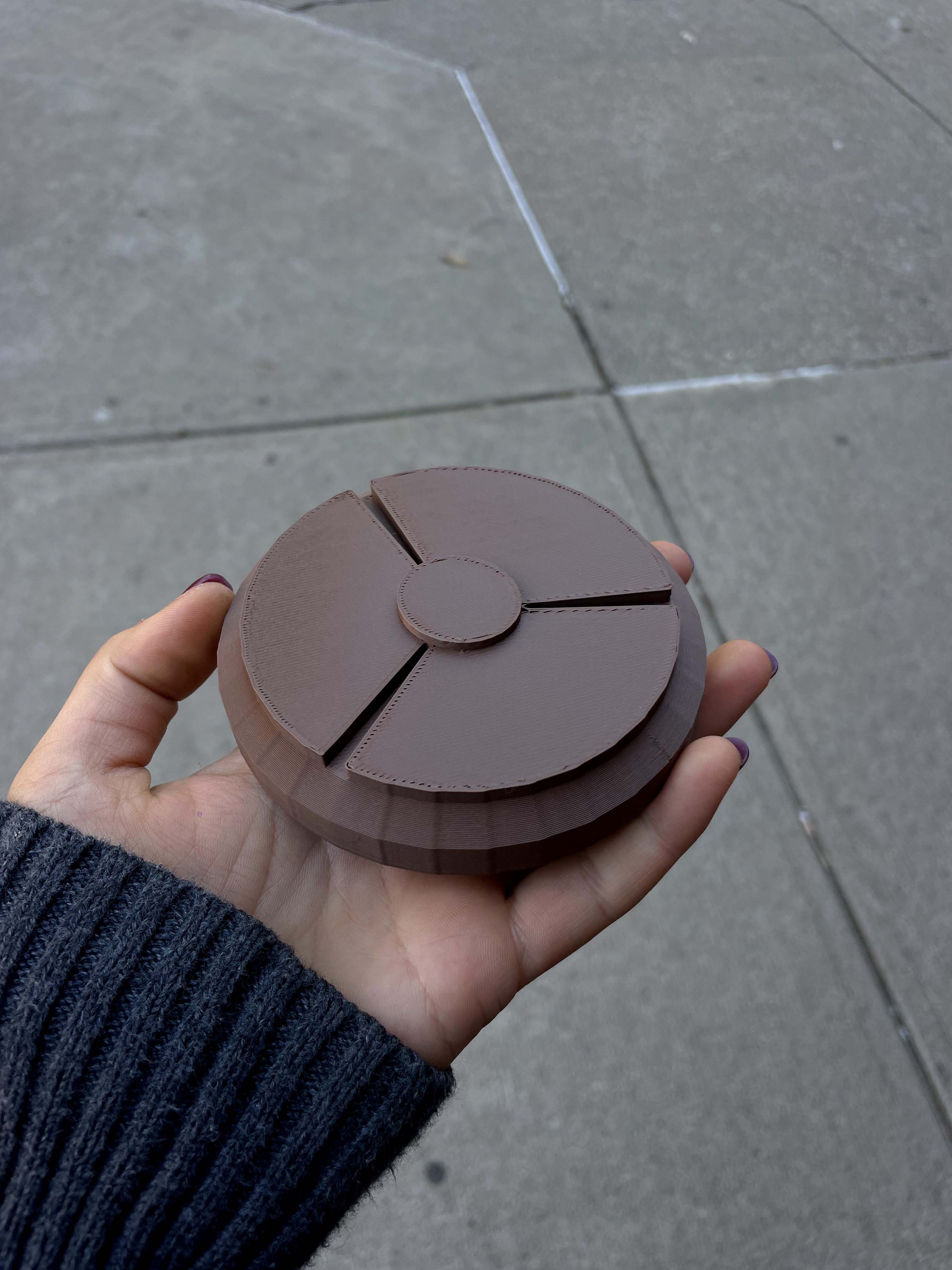 A round, brown 3D print being held in front of a concrete sidewalk background. 