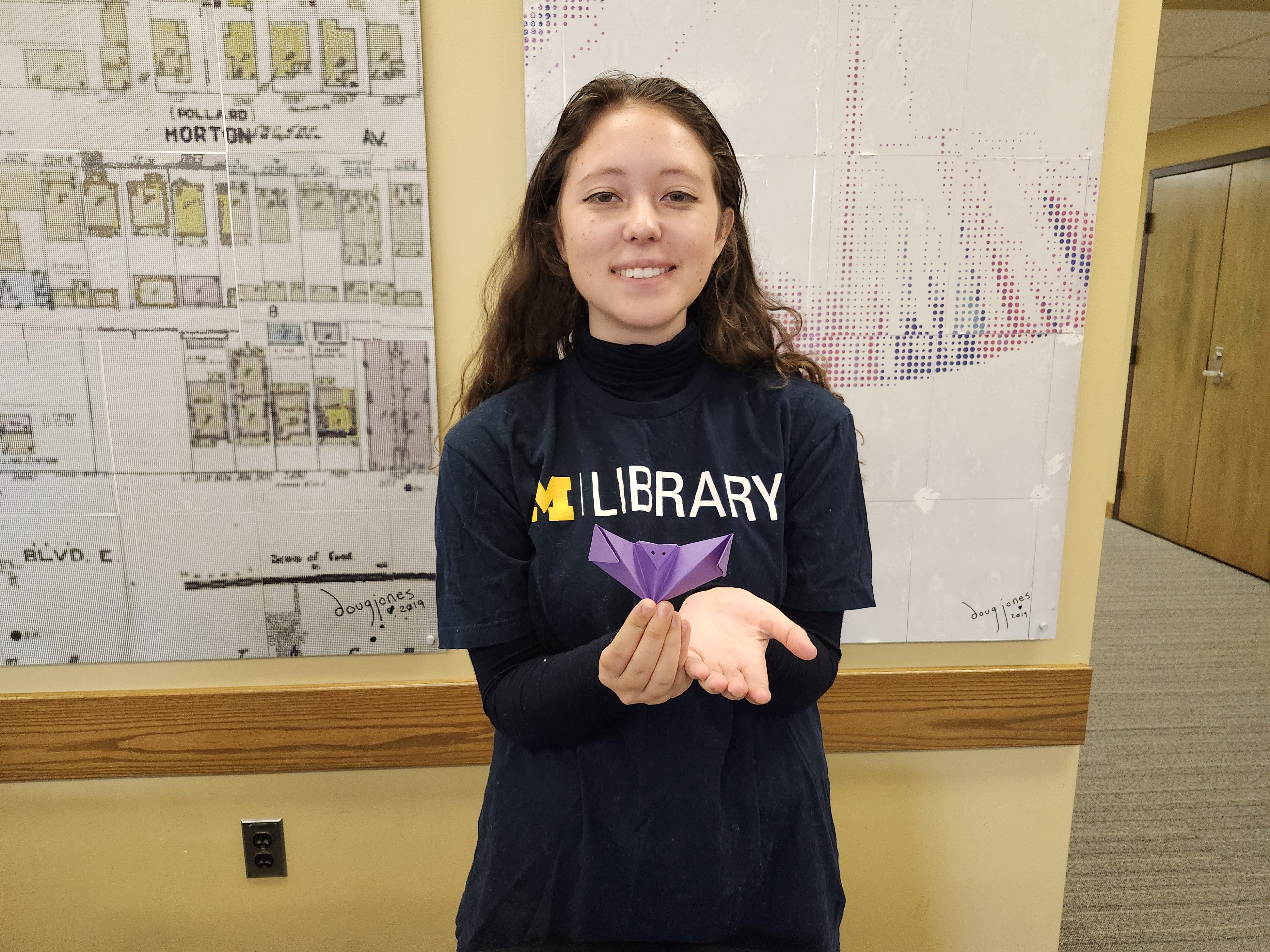 A female student, with brown hair that goes past her shoulders, holds up a purple origami bat