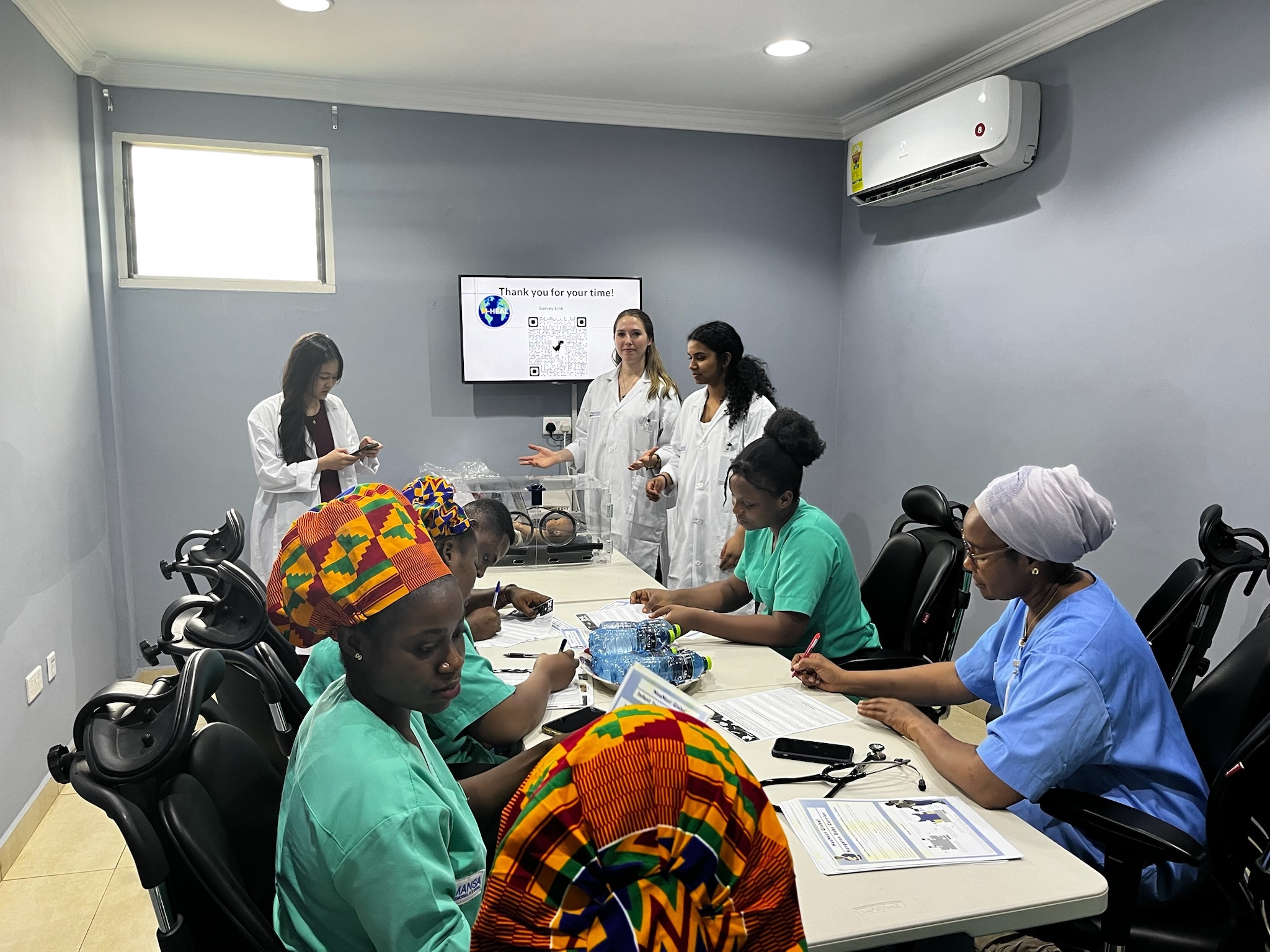 A group of students in white coats show a presentation to nurses and doctors in colorful scrubs and headwear