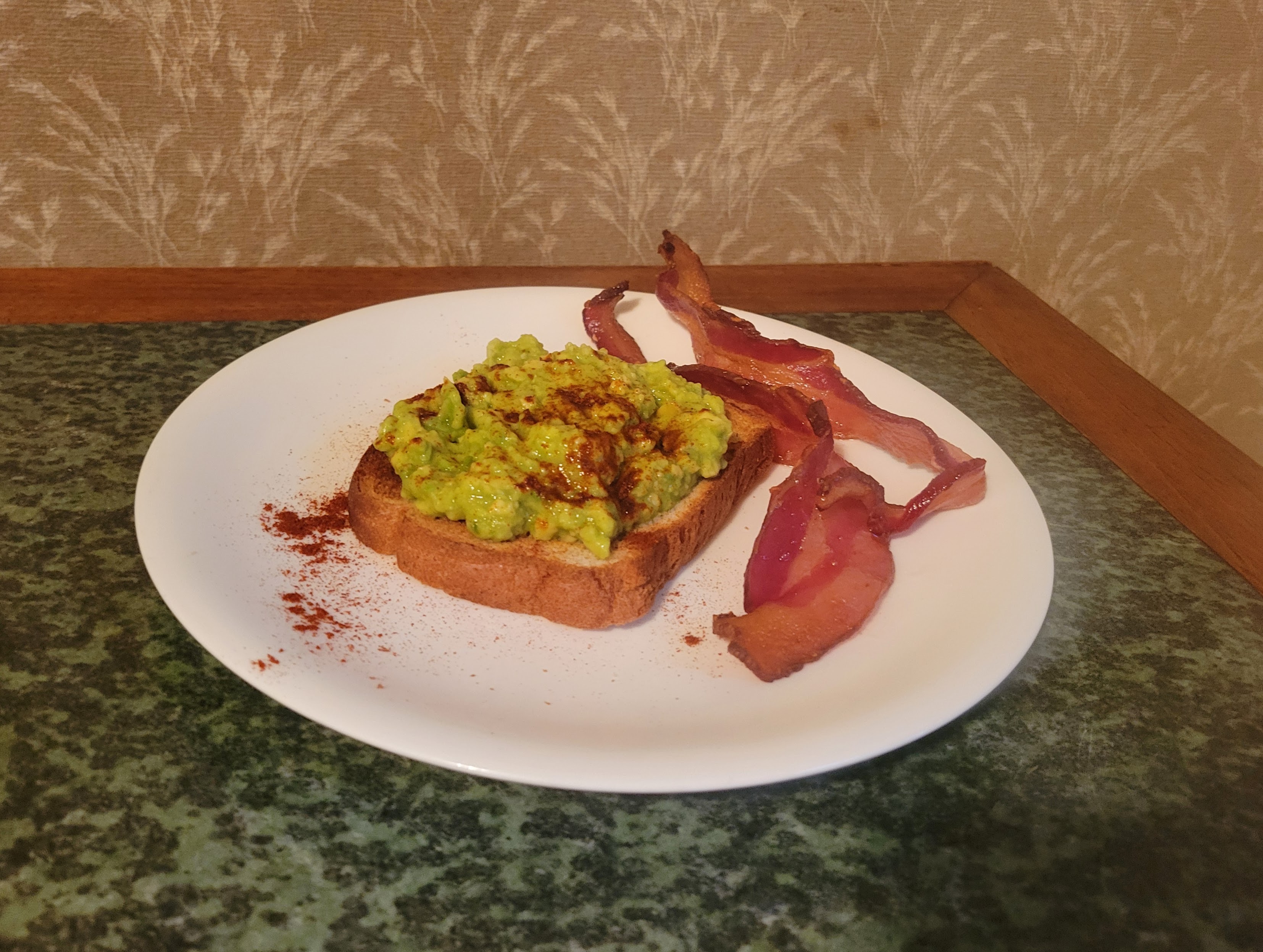 Toast piled high with mashed green avocado, sprinkled with paprika, accompanied by bacon