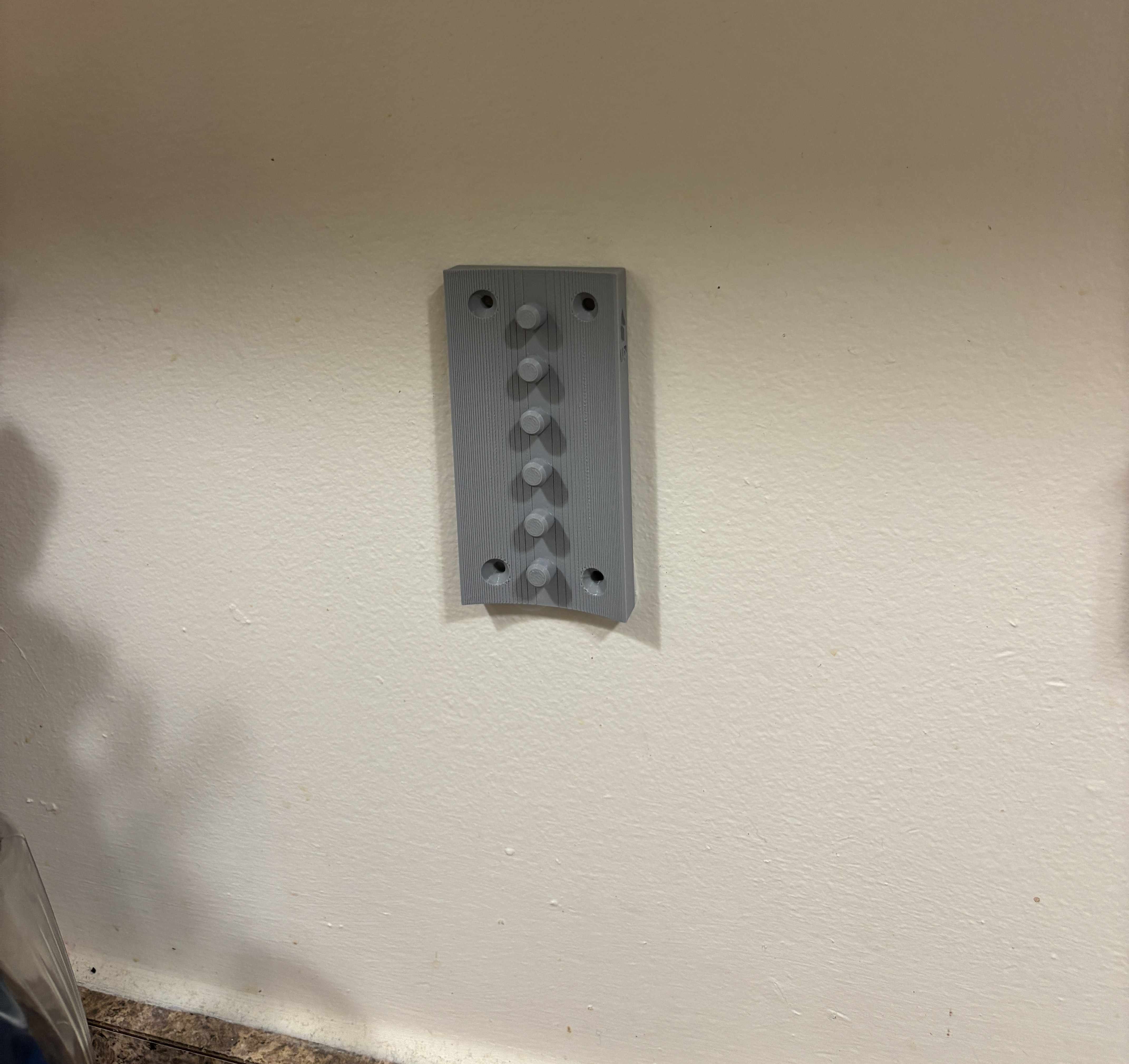 A gray 3D printed wall bracket screwed into a white wall. The bracket is a tall rectangle, with a line of small round pips down the middle.