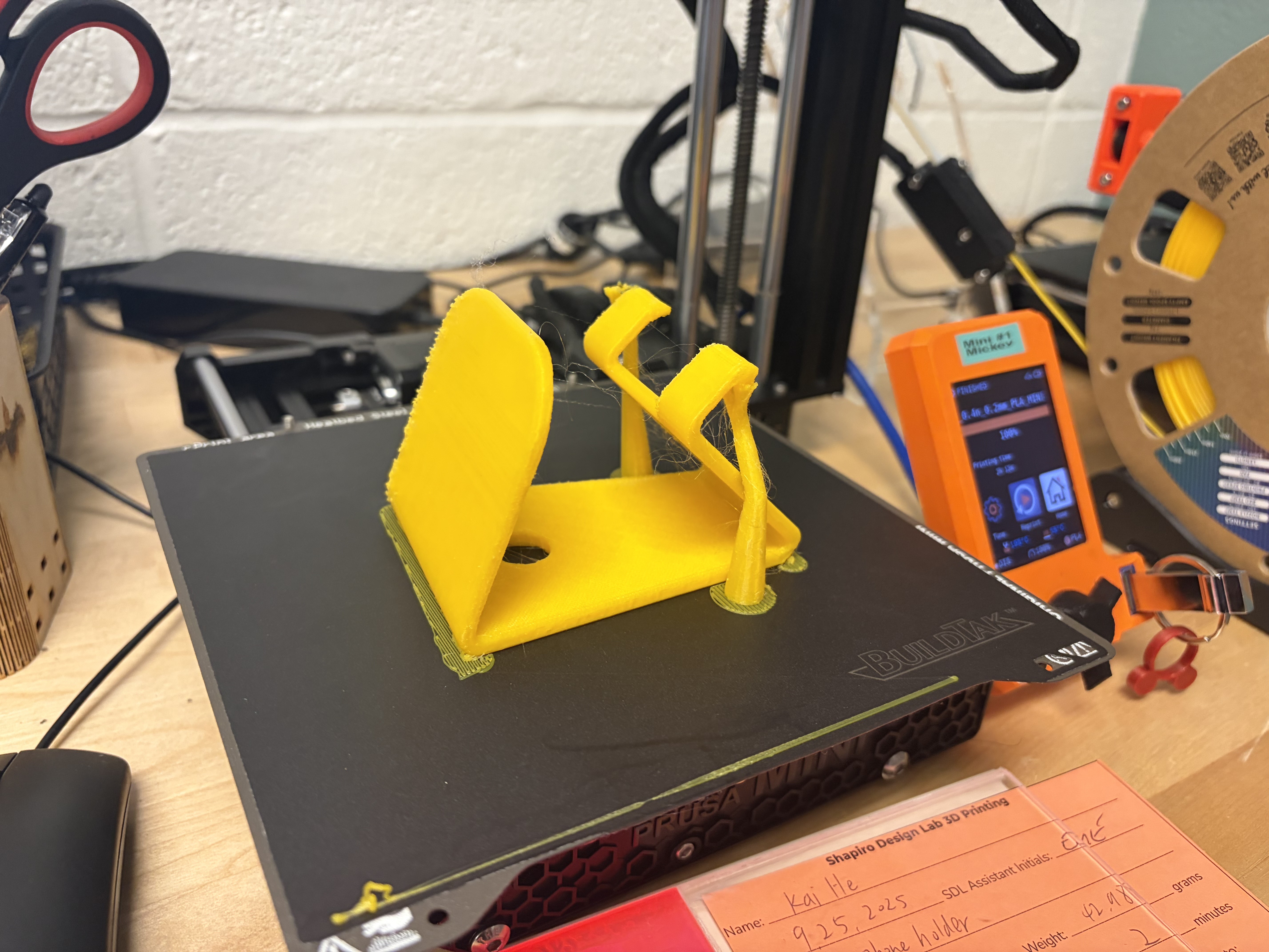 Yellow 3D printed phone holder on the 3D printer bed. The phone holder is laying on its side, and has a little brim of yellow plastic around it on the printbed.