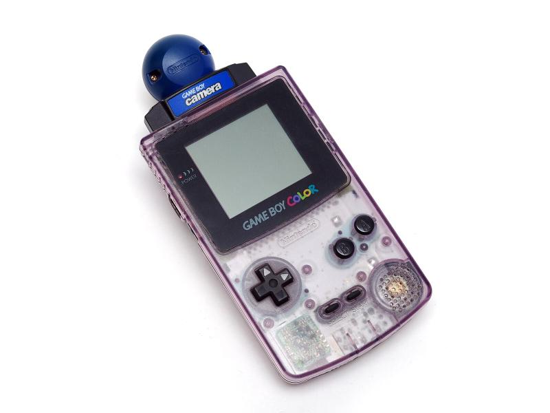 A professional picture of a Gameboy console. It is a tall rectangular console, and has a plastic, translucent silver case on the lower half of the body, and a small green pixelated screen on the upper half. Above the screen, a blue spherical camera is attached to the top of the Gameboy.