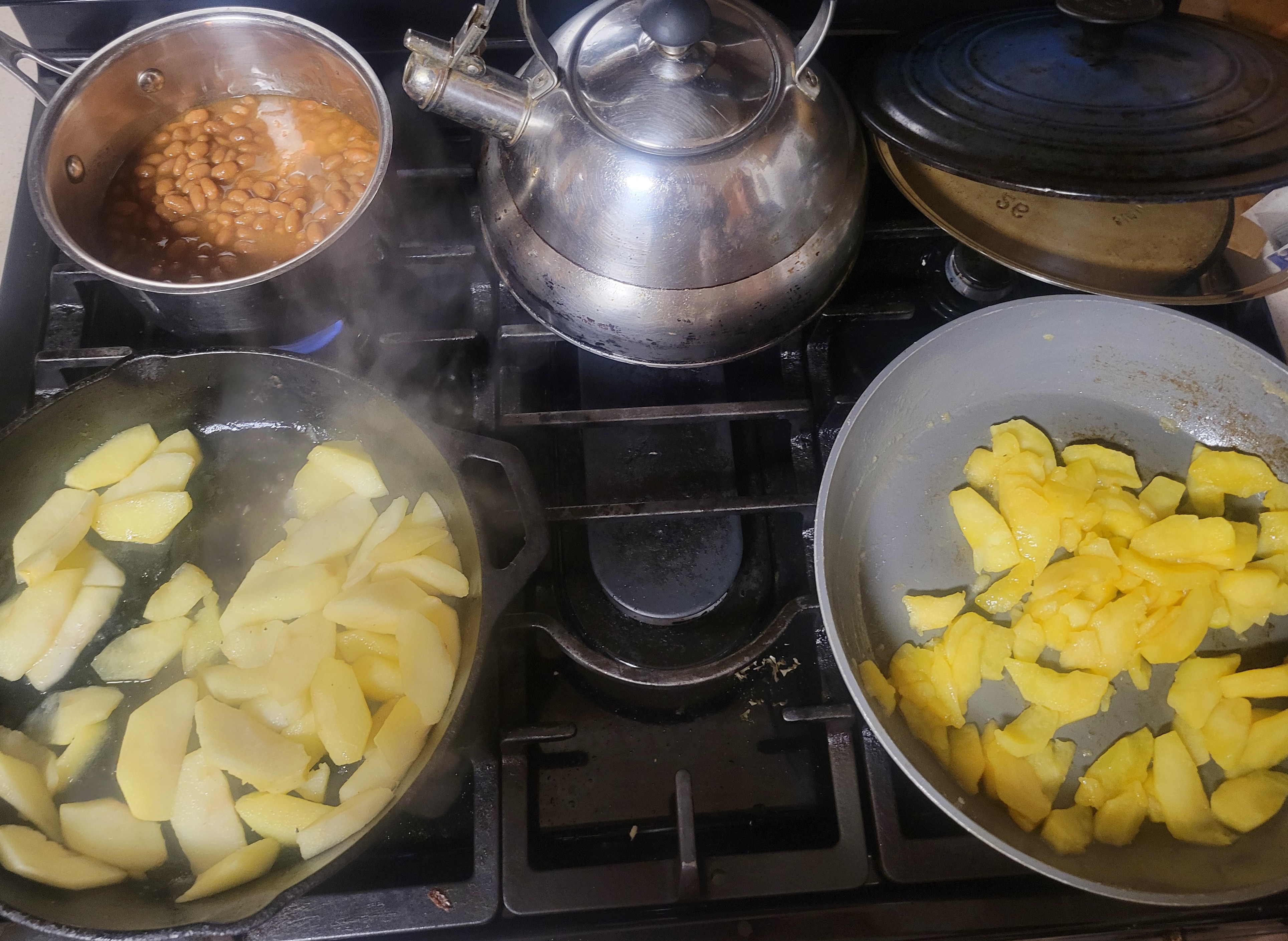 Stove top with a pan of baked beans, tea kettle and two frying pans of sliced apples. The apples on the left are pale, while those on the right are a bright yellow color