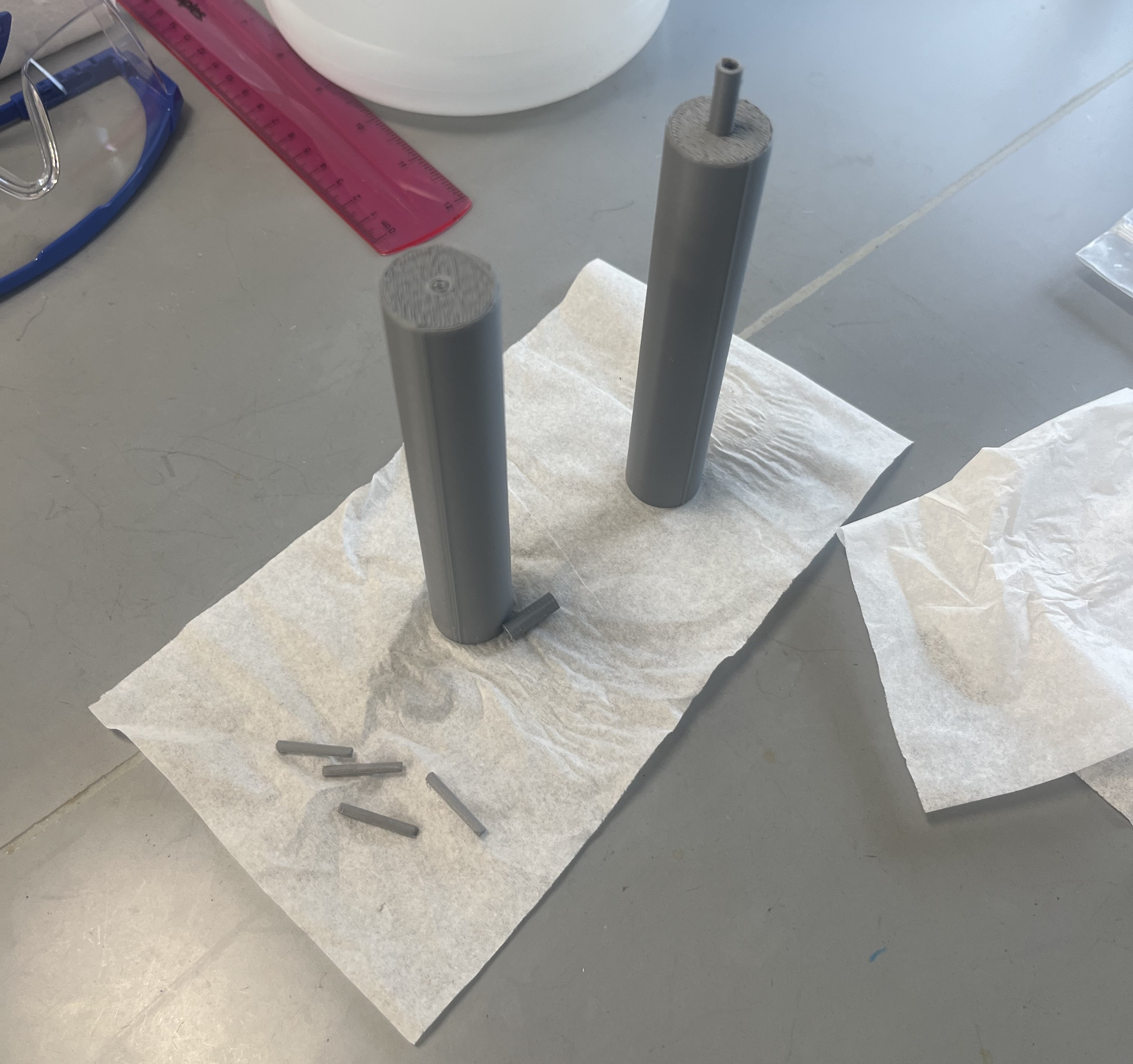 A set of gray 3D printed parts on a white napkin standing on a gray table. On the napkin are two tall cylinders, and one has a small piping end on the top. Additionally, there are four small tubes laying sideways on the end of the napkin.