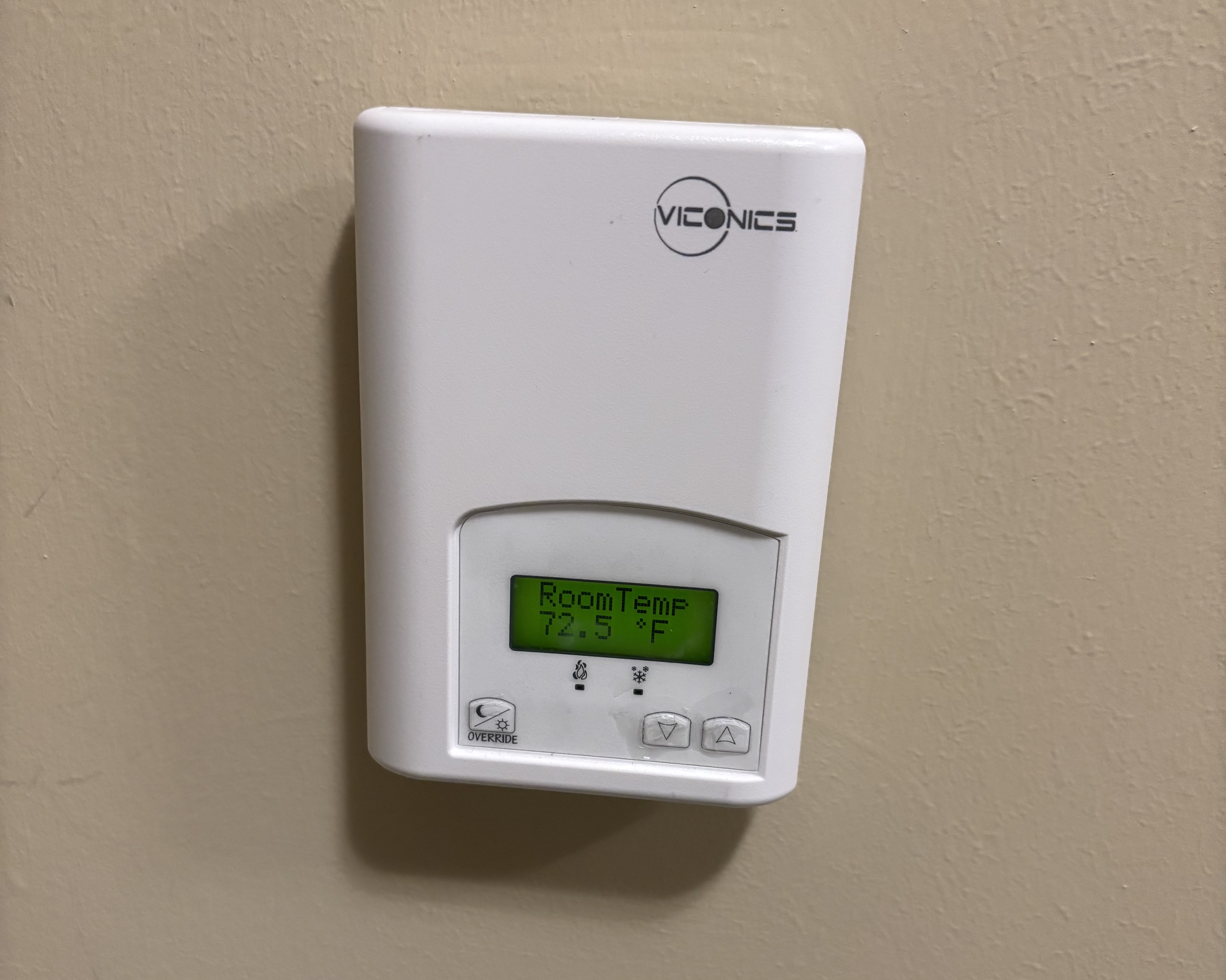 An AC control panel on a beige wall. The panel has a bright green screen near the bottom, which shows temperature, and two small buttons underneath to adjust the temperature.