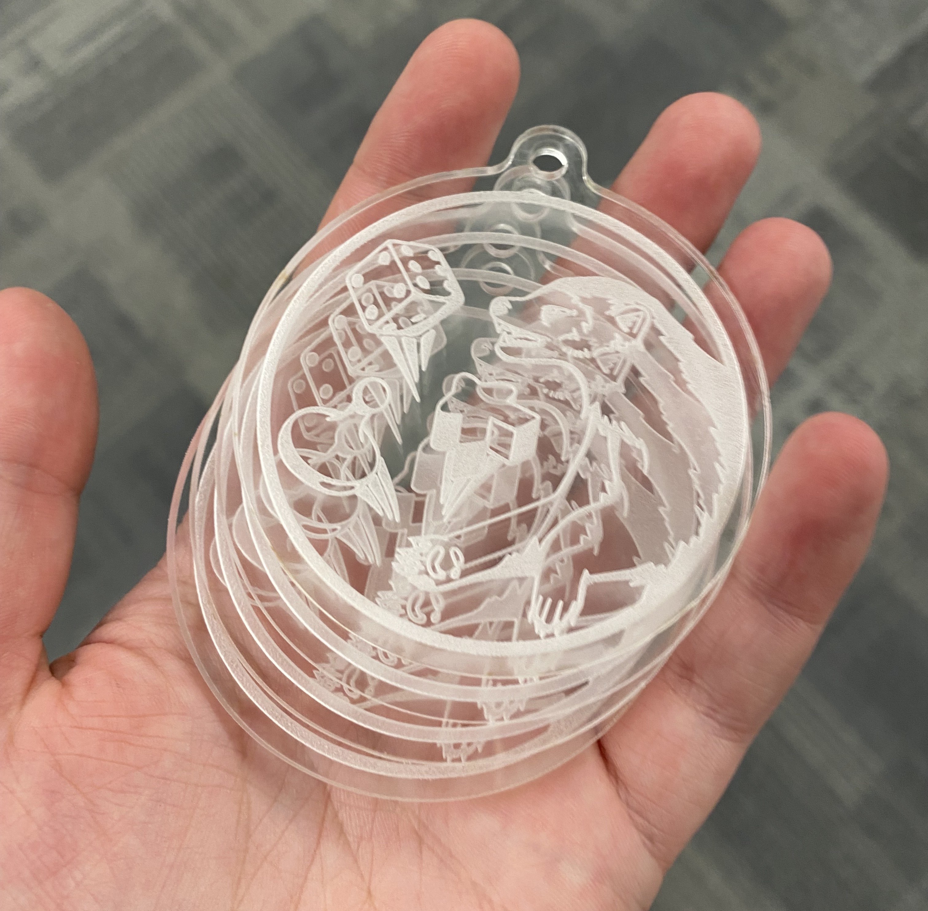 a palm-colored hand holding 4 clear acrylic, engraved, round keychains. The logo on the keychain is a realistic wolverine tossing board game pieces into the air. 