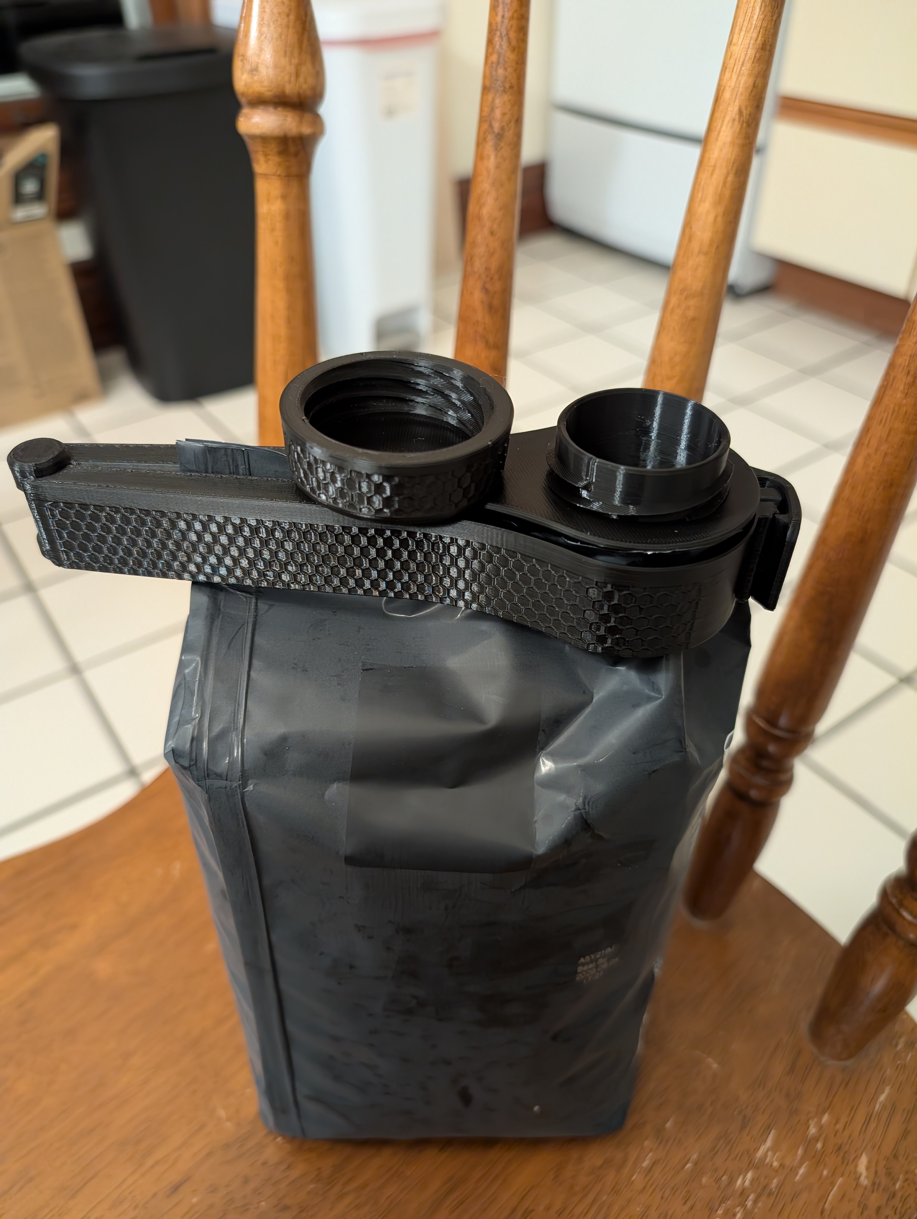 A black 3d printed bag clip on top of a coffee bag on a wooden chair. The clip has an open top and a lid that can be screwed on to close it. 