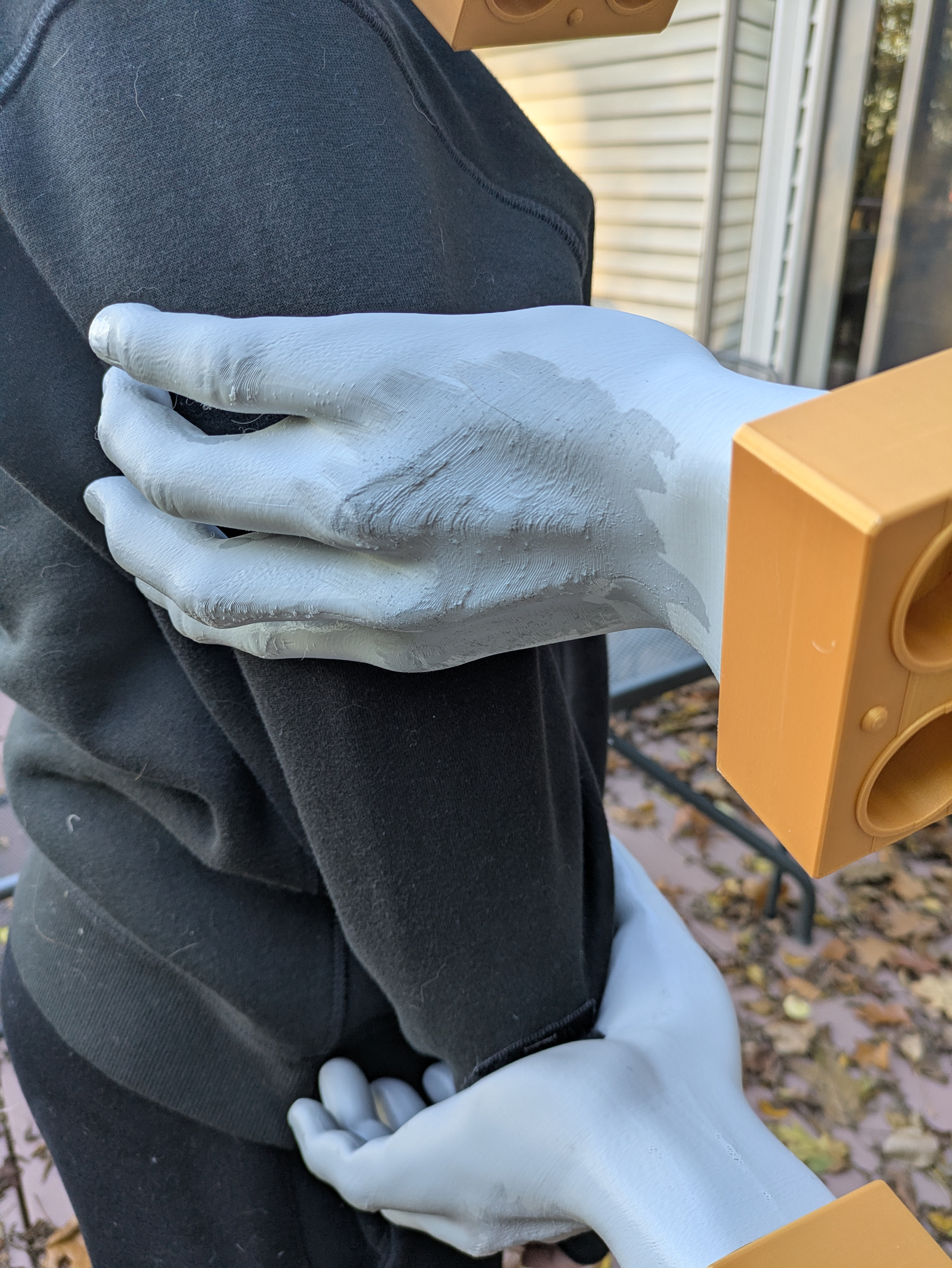 An up-close picture of a kid's arm. They are wearing a black hoodie, and two 3d printed hands - one on their upper arm, and the other on their forearm. The hands are painted gray with gold wrist connectors.
