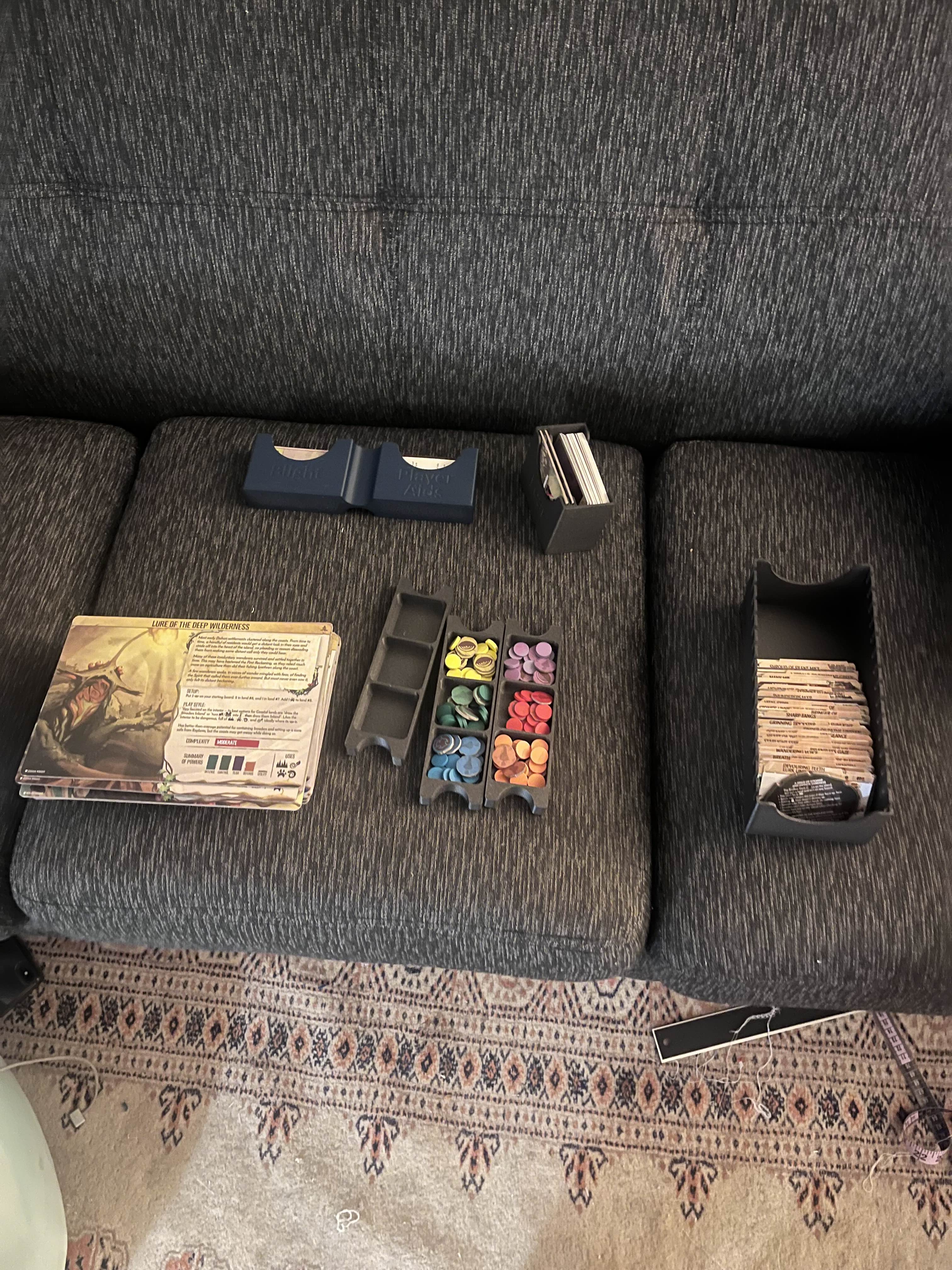 A collection of game pieces laid out on a grey couch. Included are a stack of large gameplay cards, a collection of small tokens in a 6-bin 3D printed dark grey container (sorted by color) and other smaller cards placed in small holders.  