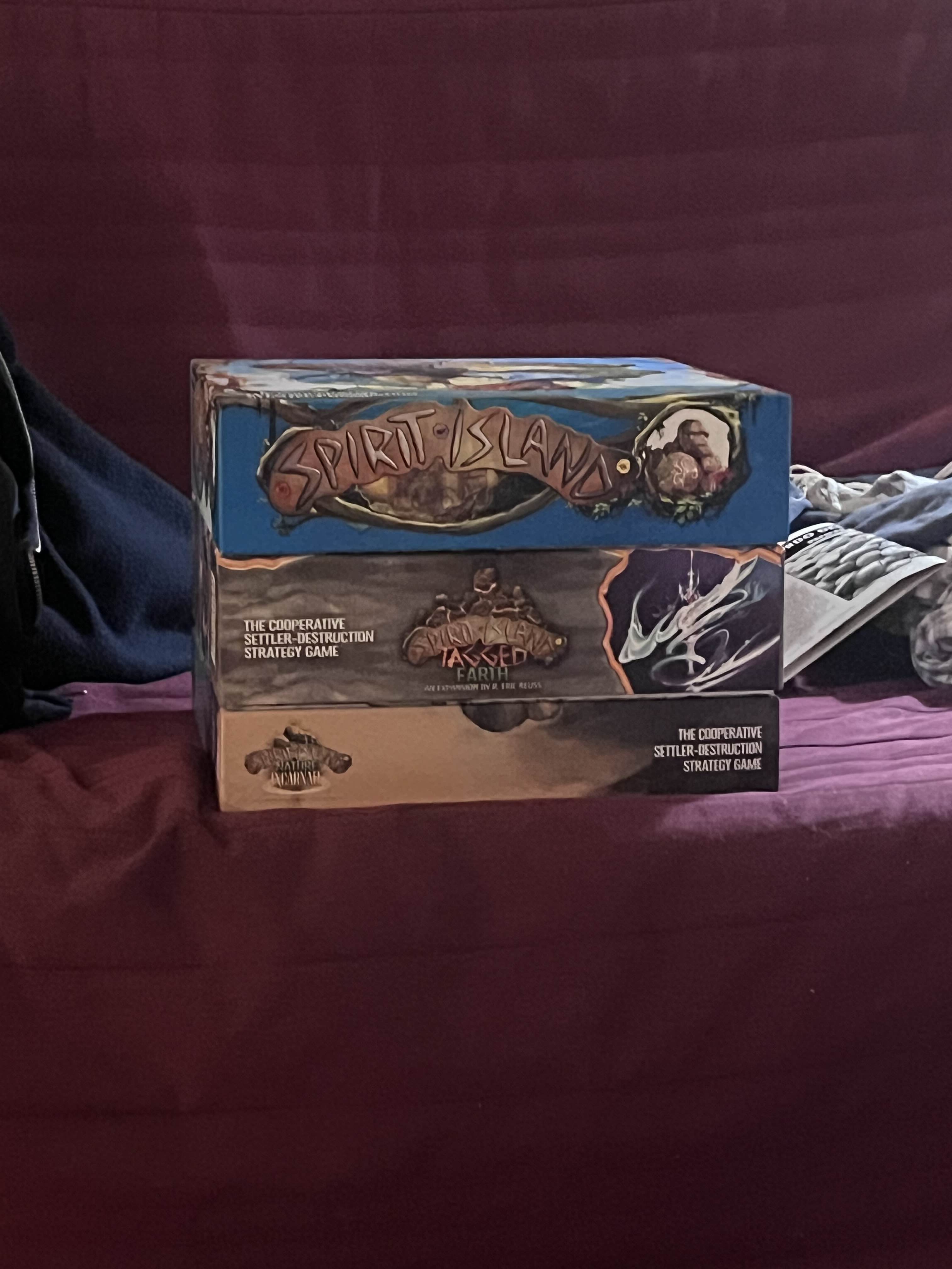 Three board game boxes stacked on top of each other on a red cushion. The top game is "Spirit Island", the middle is "Spirit Island: Jagged Earth", and the bottom one is "Spirit Island: Nature (?)". 