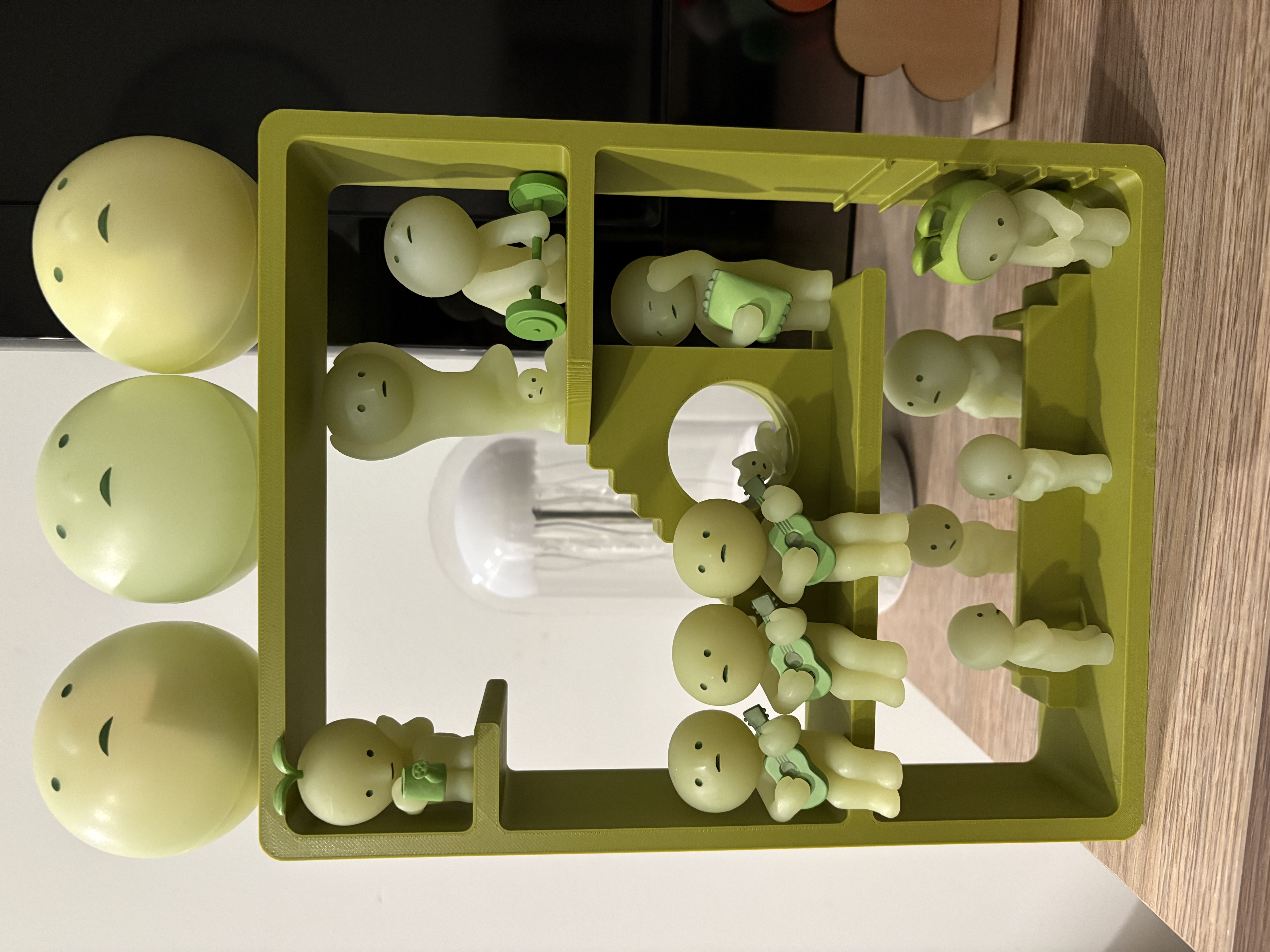 A green 3d printed cut-out of a 3-story house. There are three layers of stairs and platforms to position smiskis - small green figurines with round heads.