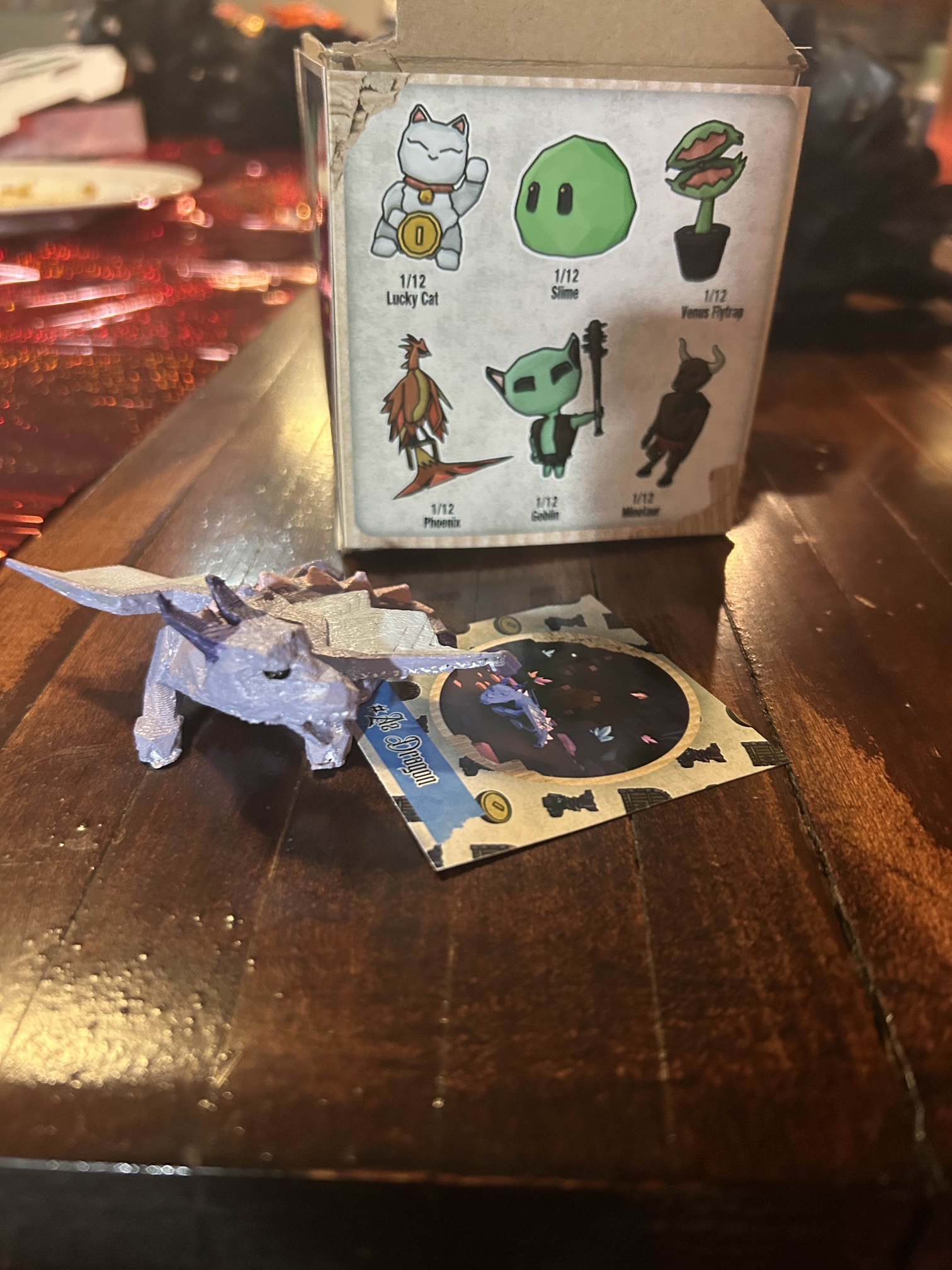 A cardboard box with six small illustrations (lucky cat, slime, venus fly trap, phoenix, goblin, and minotaur). In front of the box is a 3d printed light purple dragon with purple horns and a tan playing card with a purple swirling circle illustrated on it.