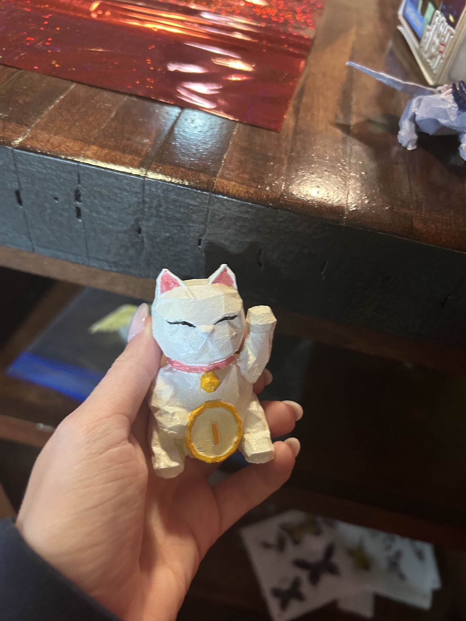 A small 3D printed figurine of a lucky cat statue. The cat is white with pink ears and collar with a gold bell, with a gold coin in front of its stomach.
