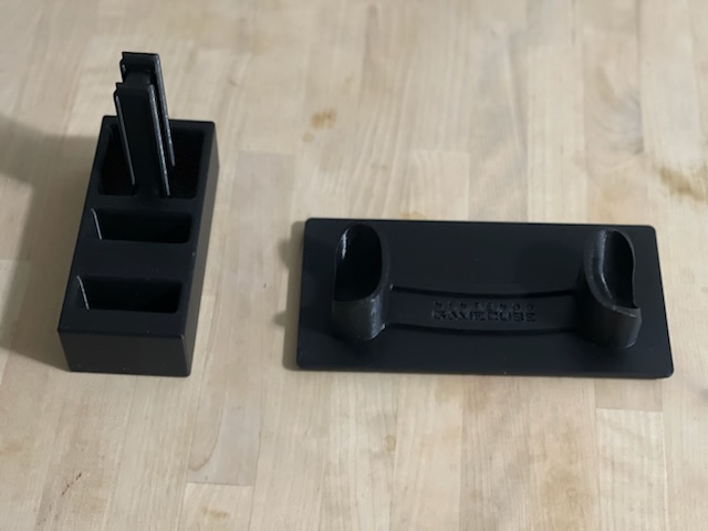 Two black 3D printed rectangular stands on a wooden table. The stands are perpendicular to each other, and one has a tall pillar in the back third, with two rectangular holes in the front two-thirds to hold the controllers. The other stand has two smaller lifts on the left and the right sides of the stand, for the gamecube controller to stand in.