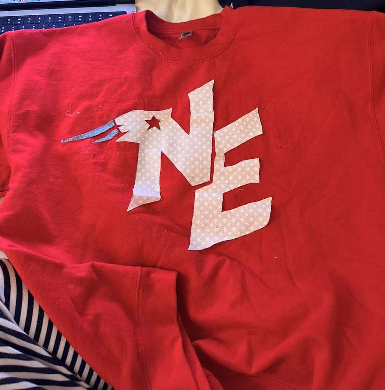 A red sweatshirt laid out on a white surface. The front of the sweatshirt has cut fabric laid out in the New England Patriots logo, but not sewed on yet. The logo consists of a white N and E, a red star in the upper left corner of the N, and from the corner are blue stripes to the left (to look like the Patriots icon).