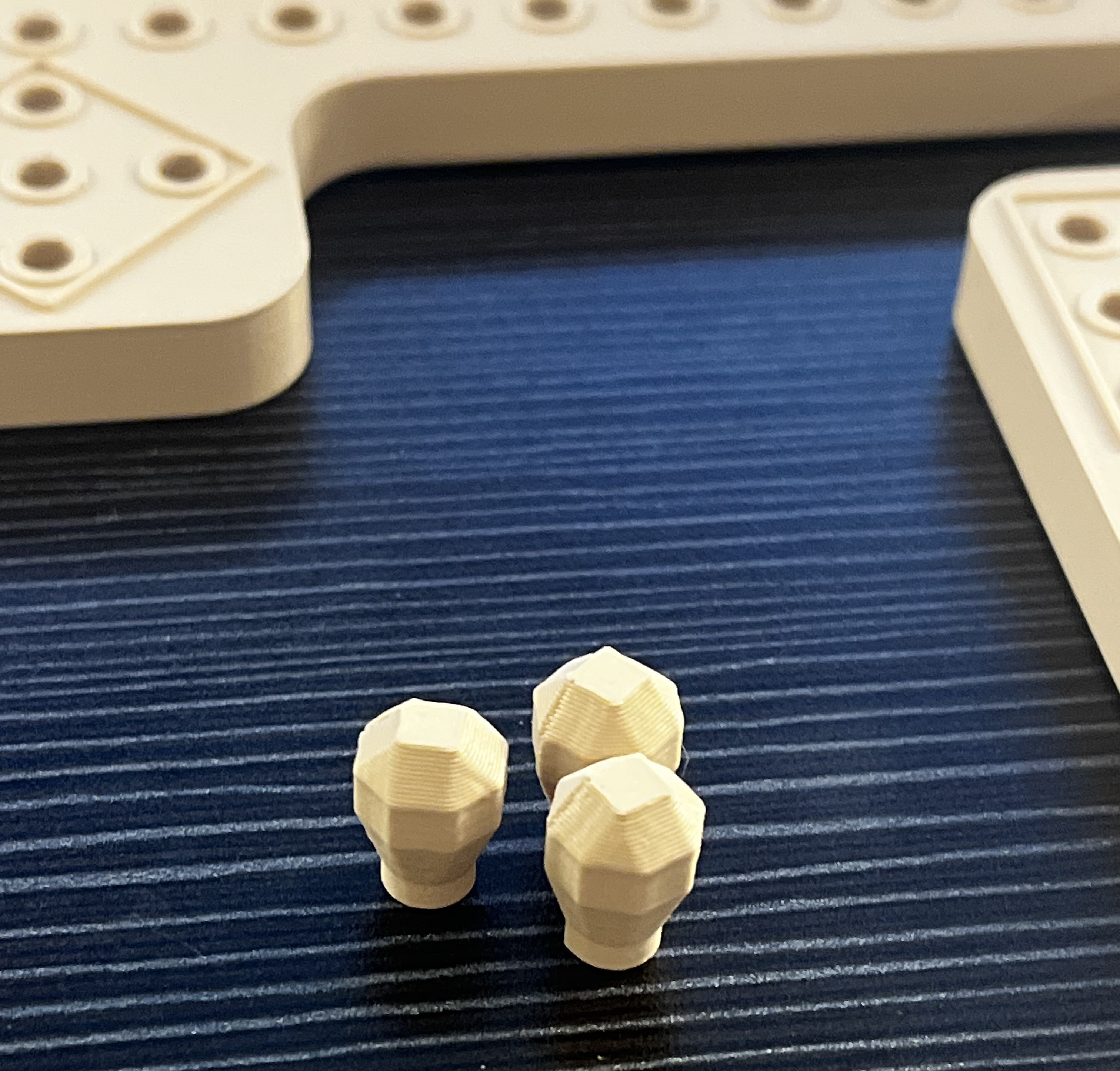 3D printed Peg and Jokers pieces on a black, pleated tabletop. The main focus of the picture are three small faceted spherical player pieces. In the background are the edges of two game board pieces, one along the top of the picture and one along the right side.