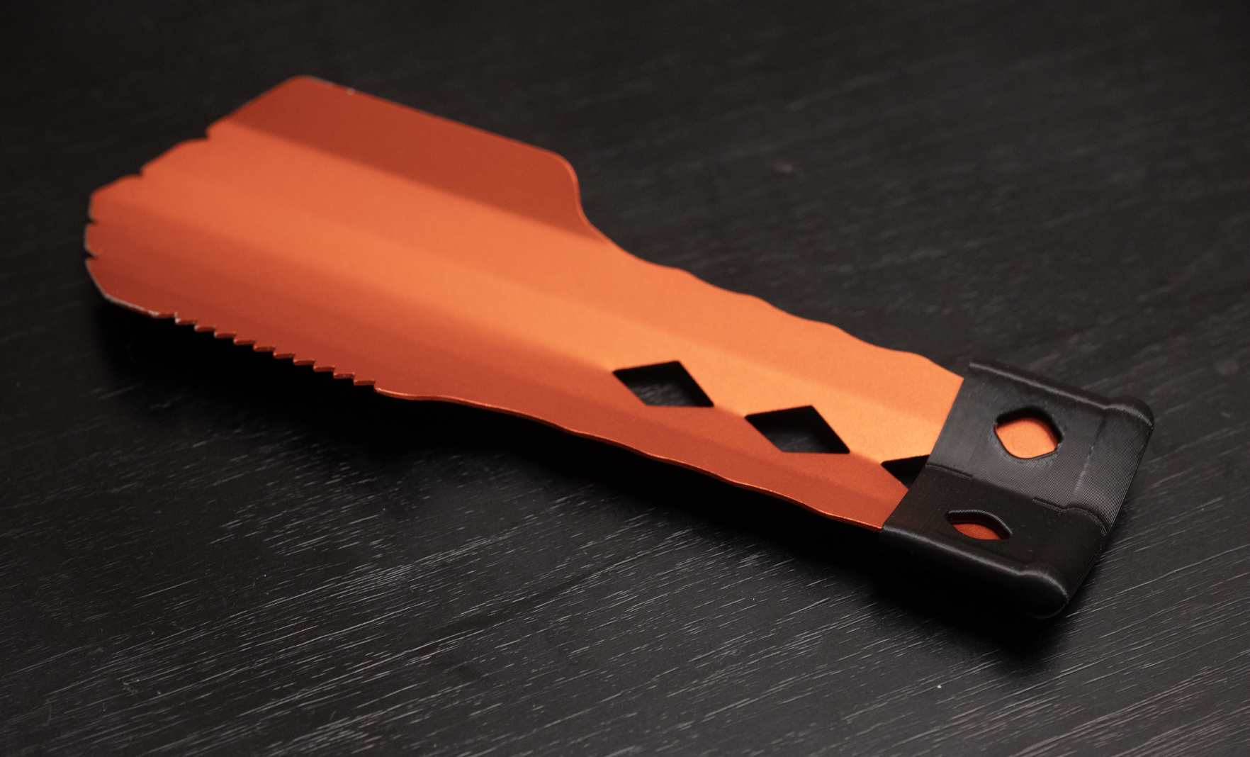 A black 3D printed cap on the end of the handle of an orange camping trowel. The cap has two circular holes where you can see the orange trowel underneath. The trowel has a blunt, slightly curved head with short teeth on the side, and down the handle are three diamond-shaped holes.