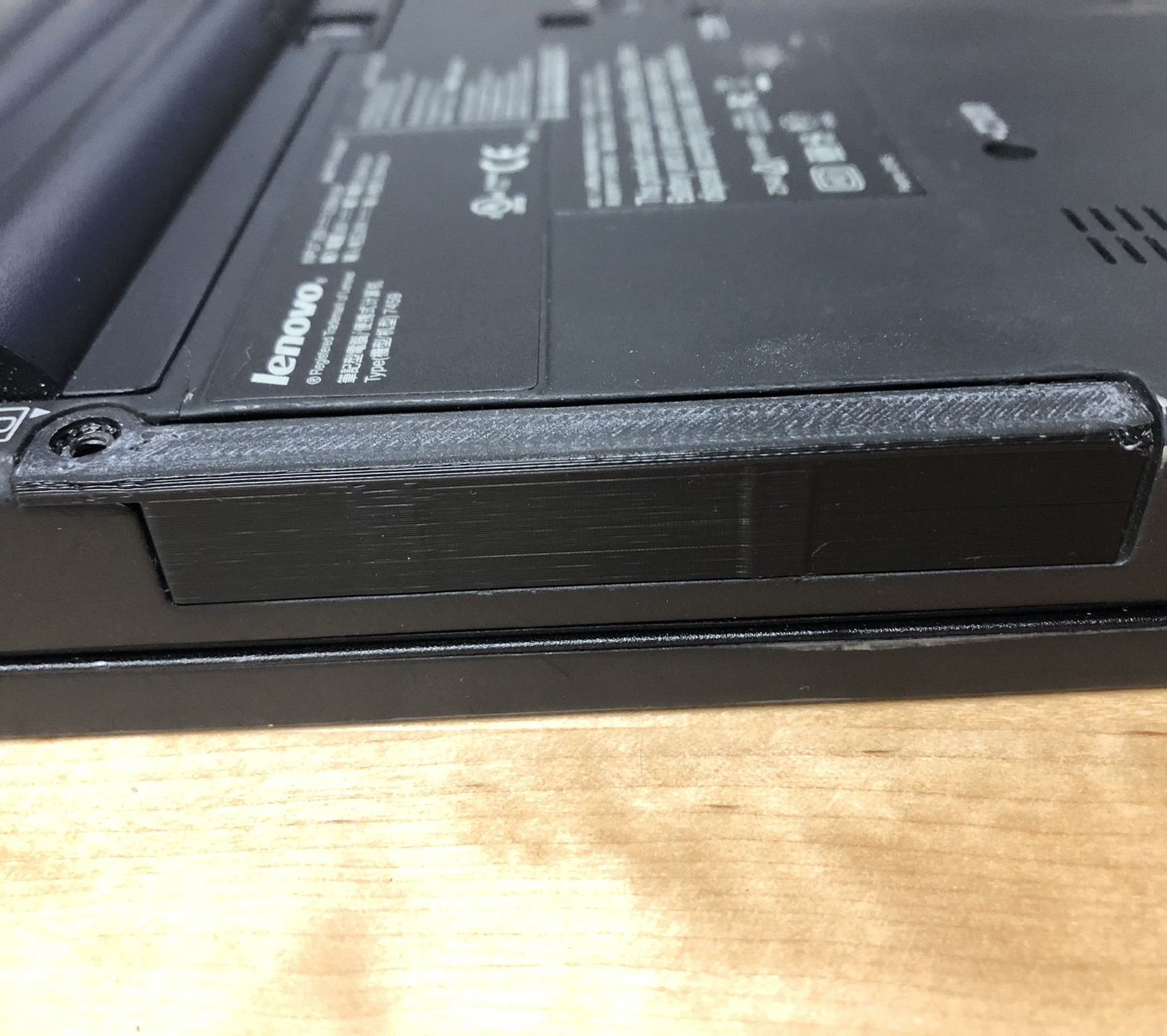 A 3d printed black, rectangular corner screwed into the edge of a laptop. The laptop is flipped upside-down, and the printed part runs along its edge.