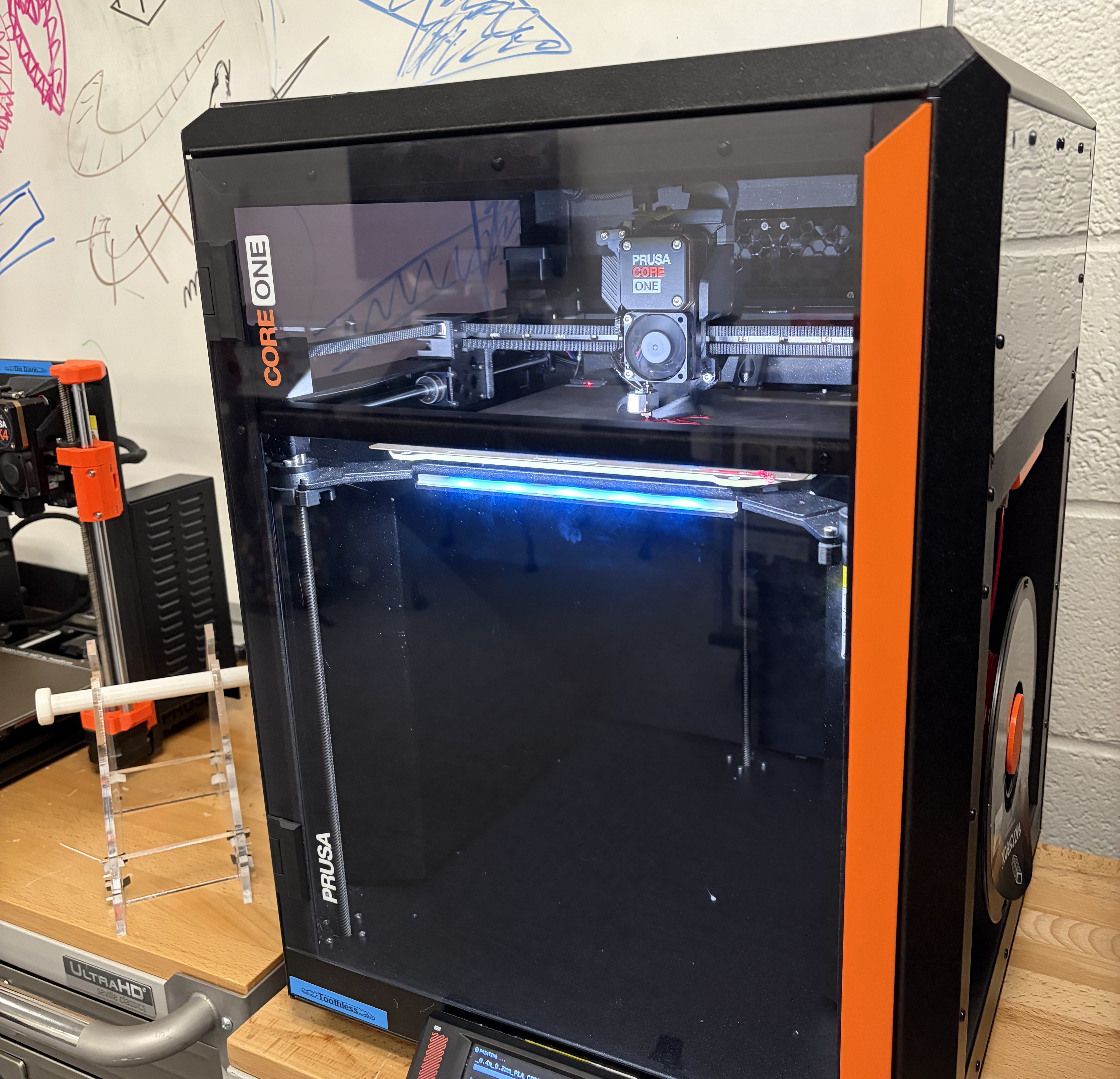 A rectangular 3D printer in the process of printing. The printer is enclosed with a plexiglass door to see inside, and the printer is blacwith orange detailing.