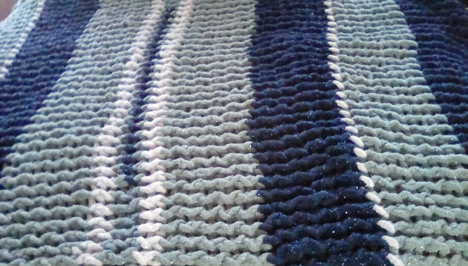 A close-up image of a completed woven blanket made out of chunky yarn. The blanket is designed to have colored rows, going up and down in this picture. From left to right, we see a dark blue columns of 5 to 6 rows of yarn, a similar column of gray yarn, a small column of white-blue-white (about 2 rows each), followed by gray-blue alternating columns (about 6 rows each)