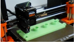 A close-up of a 3D printer printing a puzzle piece of a loom. The print is lime green, on a brown build plate on a black and orange printer.