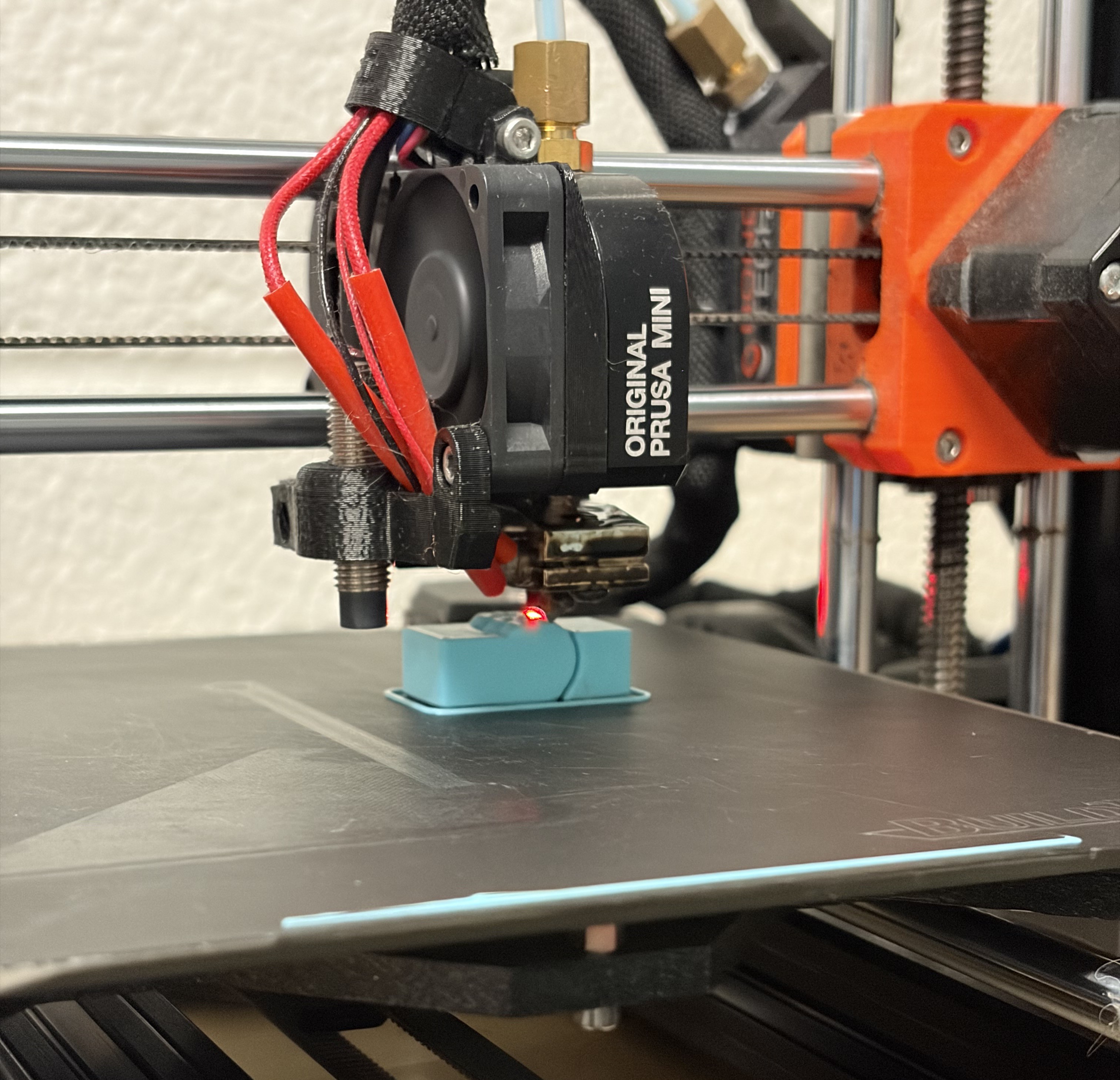 A light blue rectangular object being printed on a 3D printer. The printing plate and the printer head is black, and the arm the printer head is attached to is metallic with black and orange accents. On the side of the printer head it reads, "ORIGINAL PRUSA MINI". At the front of the print plate is a line of light blue filament.