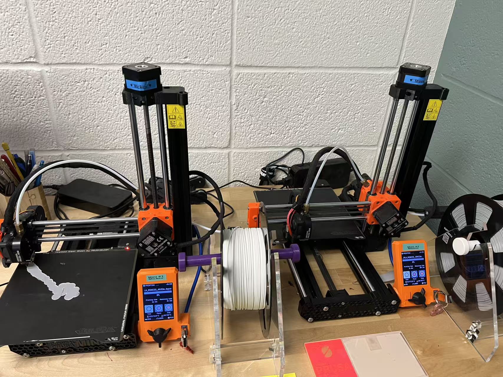 Two Prusa Mini 3D printers on a wooden table, in front of a white cinderblock wall. The printers are side by side, and each one is back with orange accents. Both printers have a roll of filament attached (white on the left, gray on the right printer) and both are actively printing.