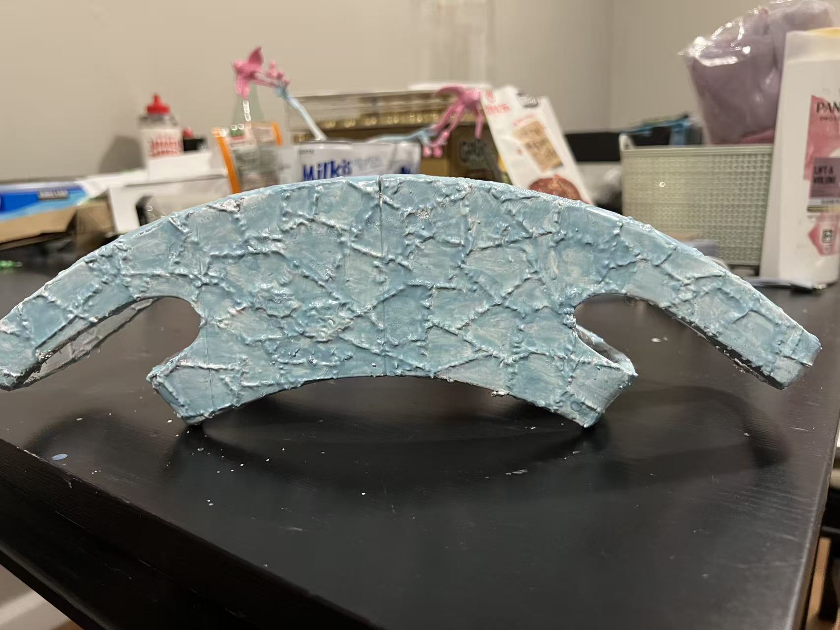 A painted traditional ceremonial crown. The crown is a large rectangular shape, with the edges of the rectangle curving down to look semicircular from the front; the top of the crown is much longer than the bottom. There is various detailing across the entire crown, and it is painted blue-gray and gray around the entire crown. The crown is on a black tabletop, and in the background various small, out-of-focus trinkets and pens are visible.