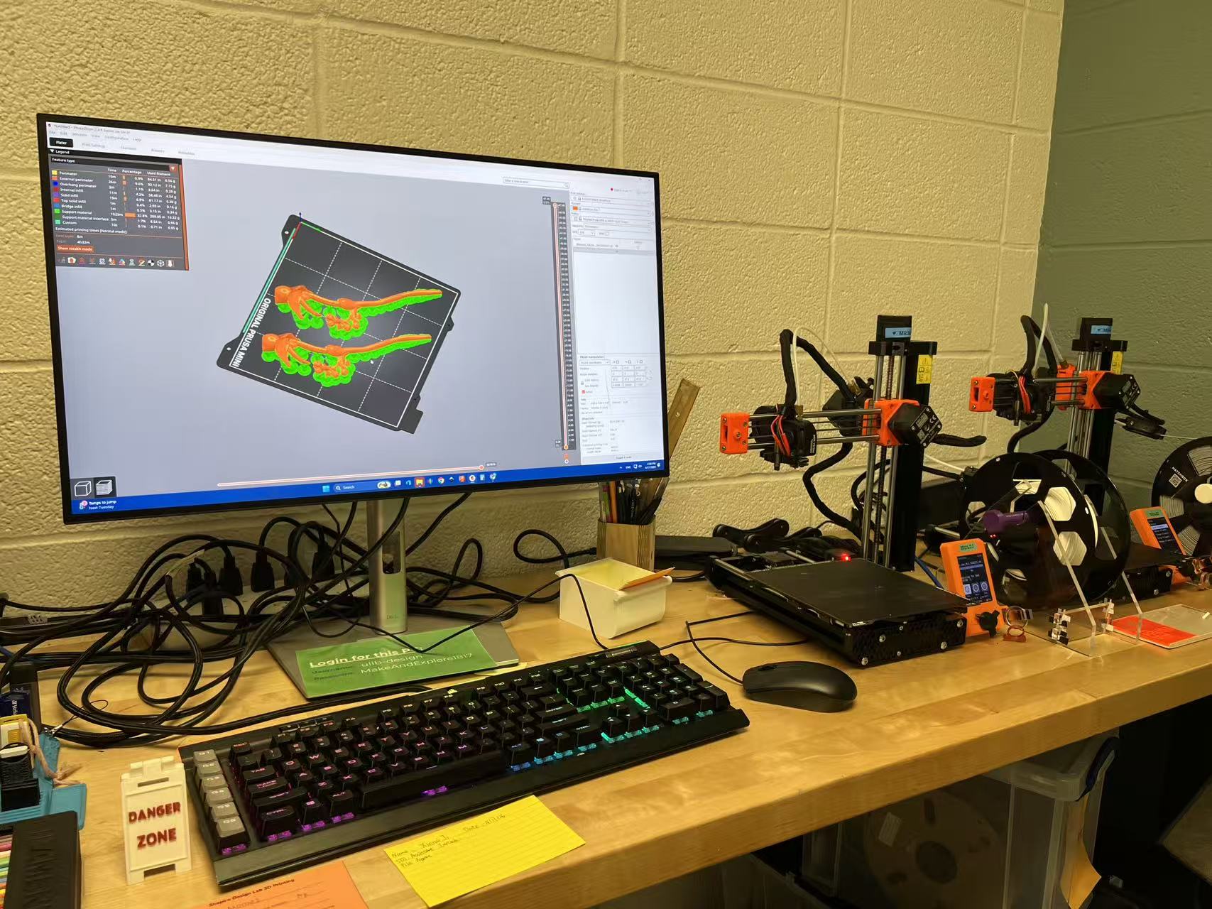 A desktop computer and two Prusa Mini 3D printers, from left to right, on a wooden table in front of a white cinderblock wall. The computer screen shows a 3D print slicer of two flower hairpins on a print bed; the prints are colored in orange, and the supports are in green. The two 3d printers are black, with orange accents, and each has a roll of filament attached. The way the picture is taken the corner of the room is visible behind the second 3D printer on the far right, and the other wall is gray-green.