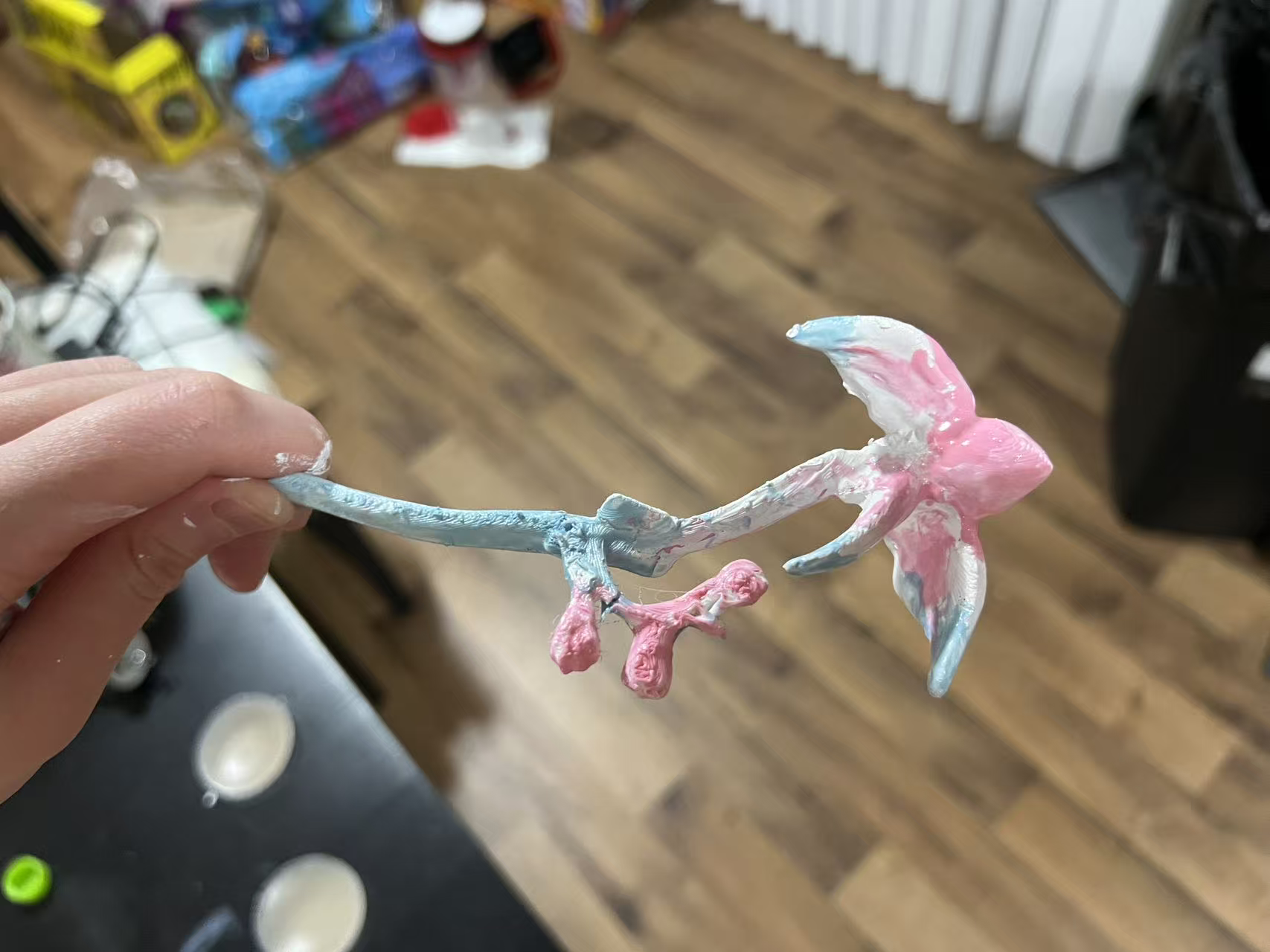 A white hand holding a painted flower hairpin in front of a hardwood floor. The stick of the pin is painted blue, and the flower and buds are painted white and pink. In the background, an out-of-focus table with plates is visible in the background.