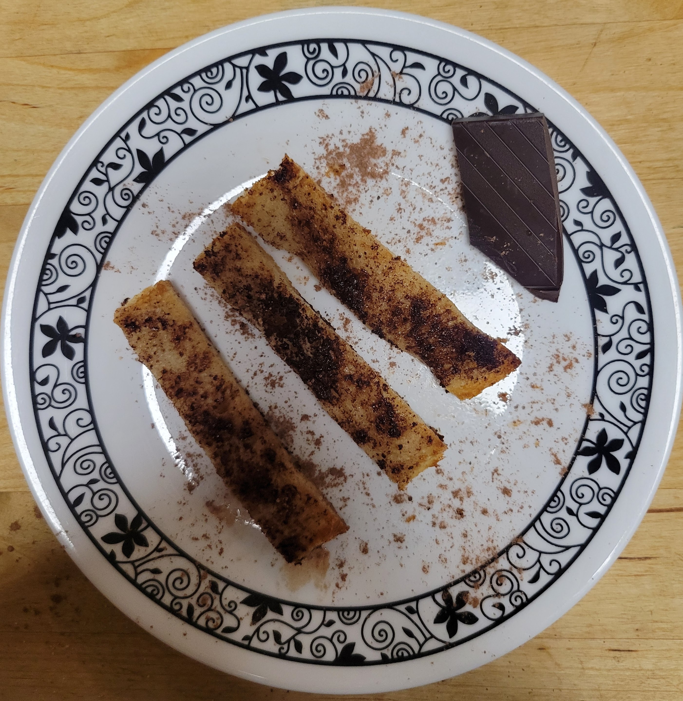 Three rectangular toast sticks with brown cocoa powder on a white plate with a piece of chocolate on the side