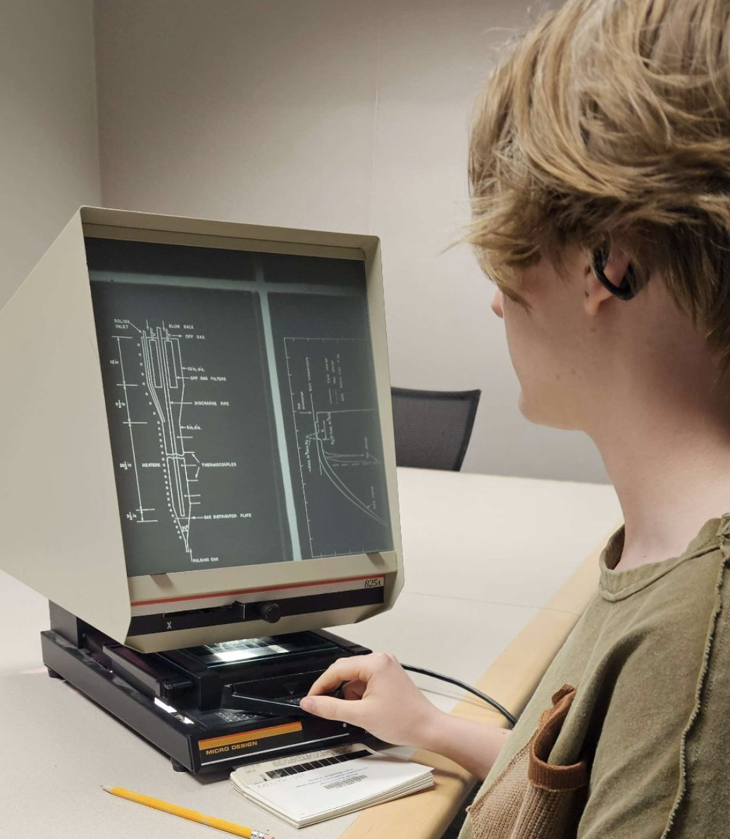 Author Atlas Fracasso using a microfiche reader to examine a technical report stored on a microfiche during the cataloging process.