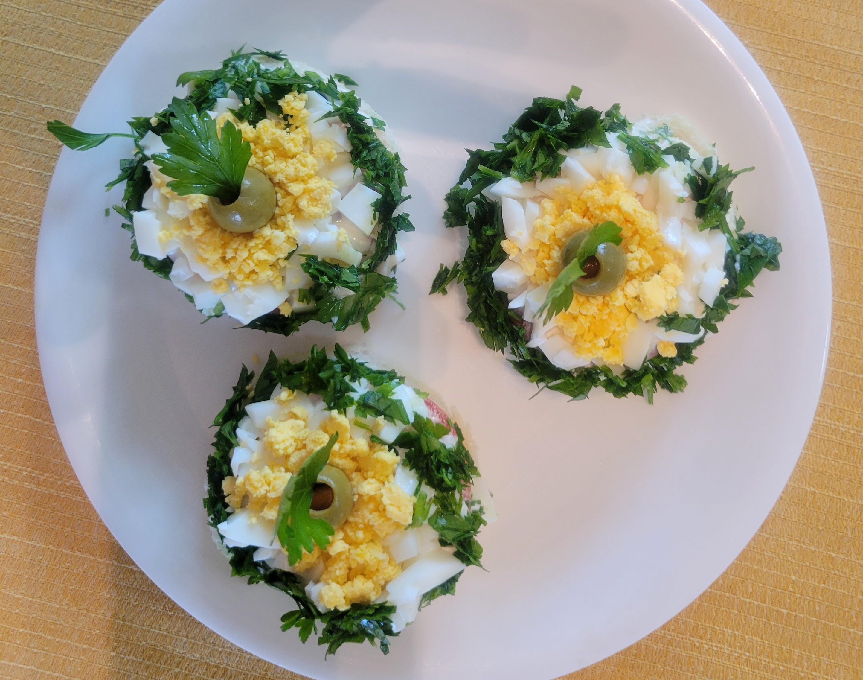 Another photograph of the tomato canape appetizer. This time a view from above showing the circles of green parsley, white egg white, yellow egg yolk, and green olive