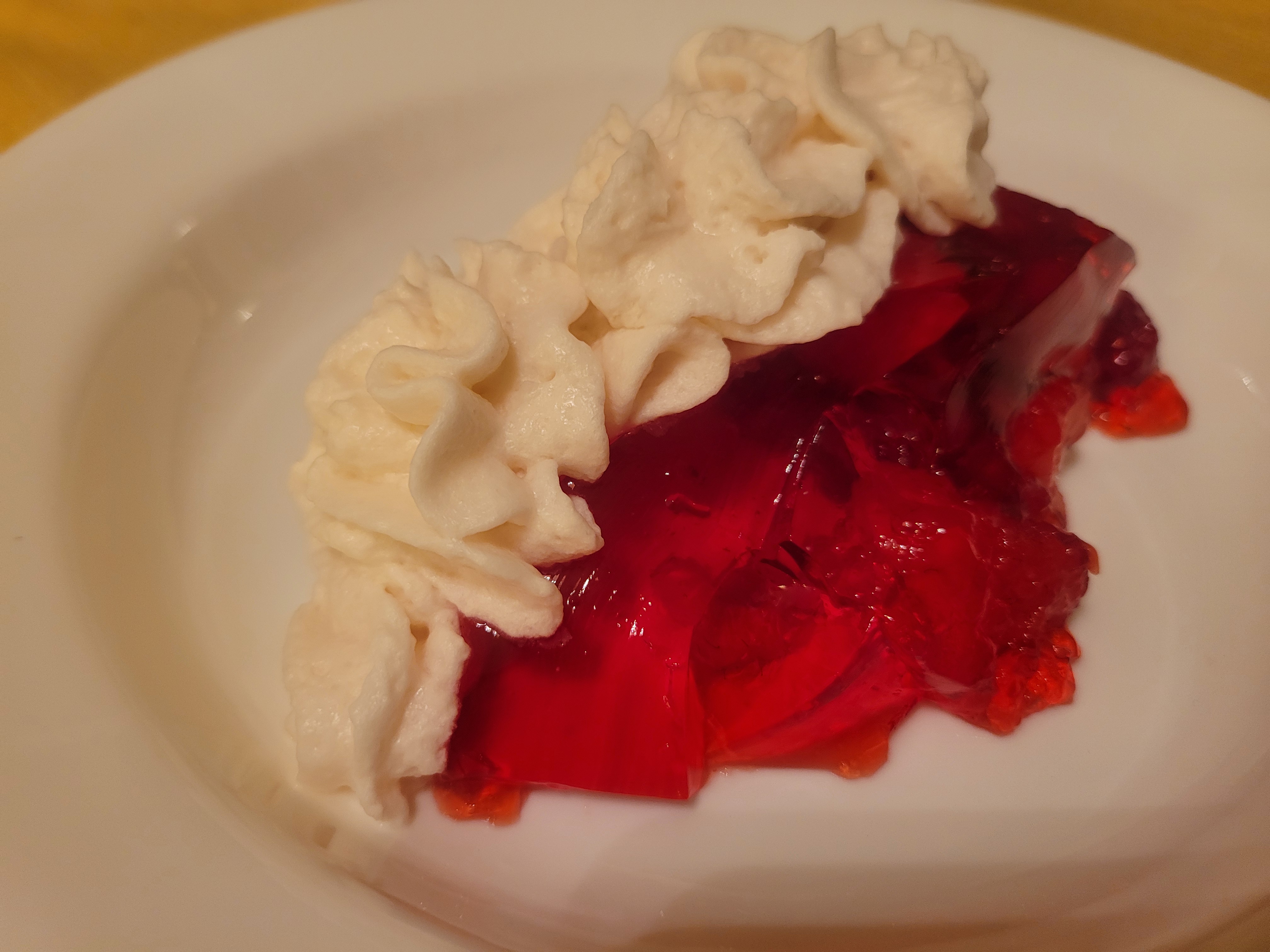 An individual slice of red jell-o on a white plate, with a line of whipping cream running along the top