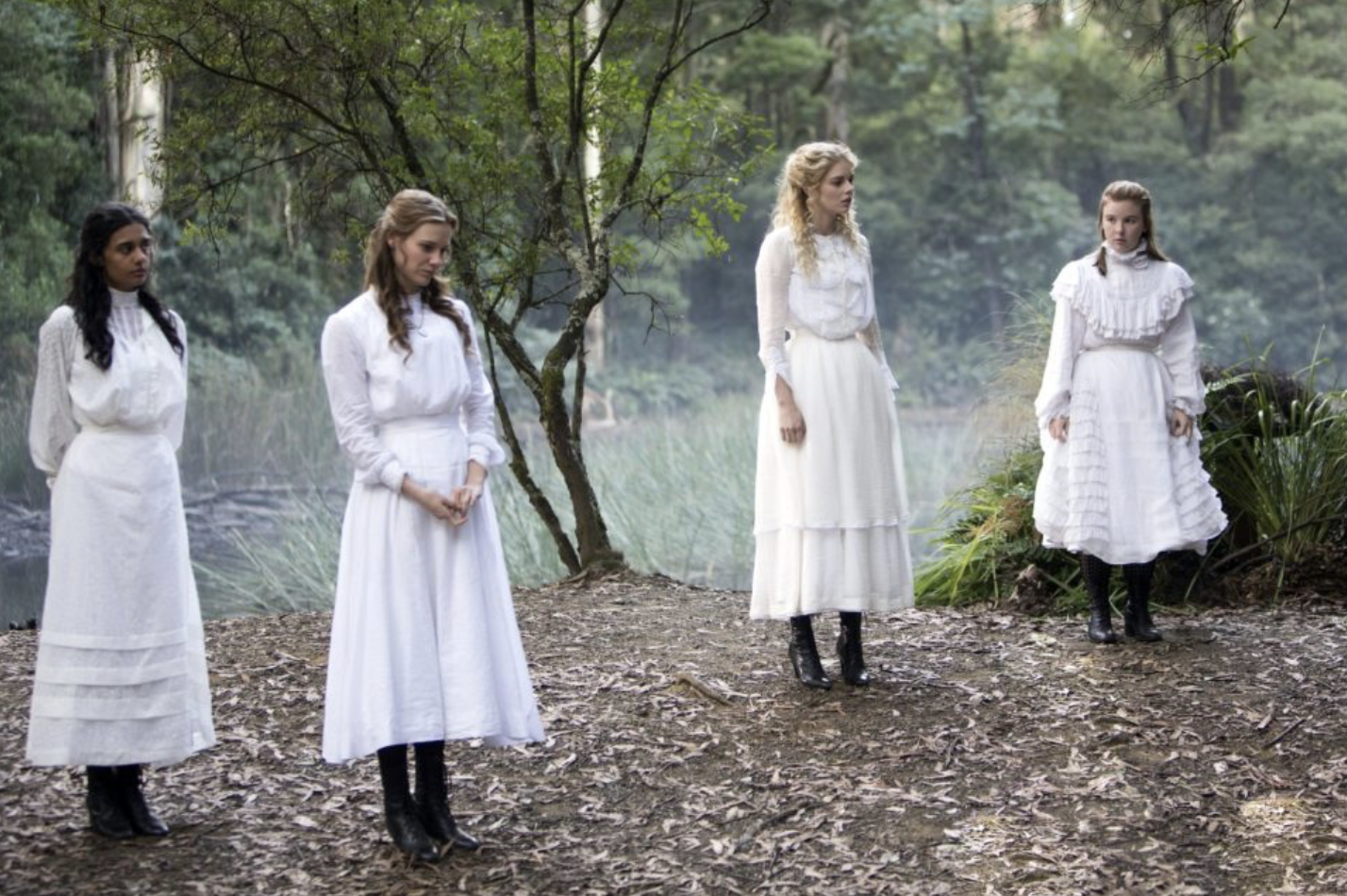 Four young women wearing long white dresses stand in the woods.