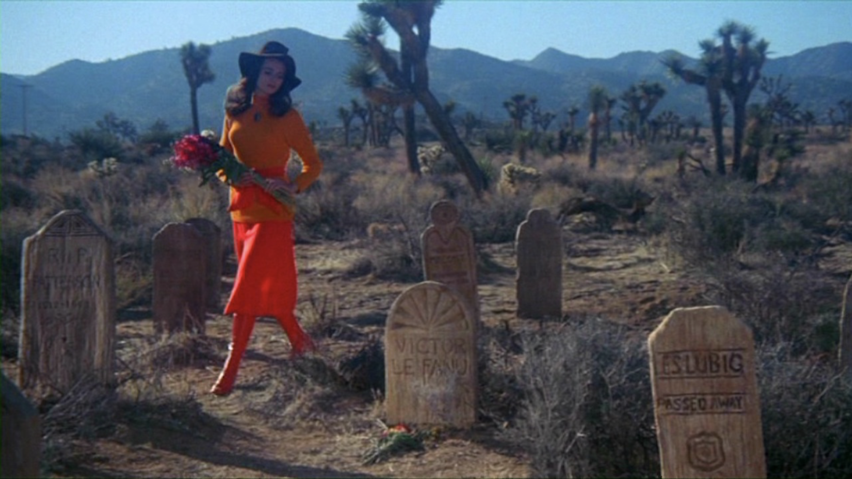 A woman wearing colorful red and orange clothing with a large black hat walks through a graveyard holding a bouquet of red roses.