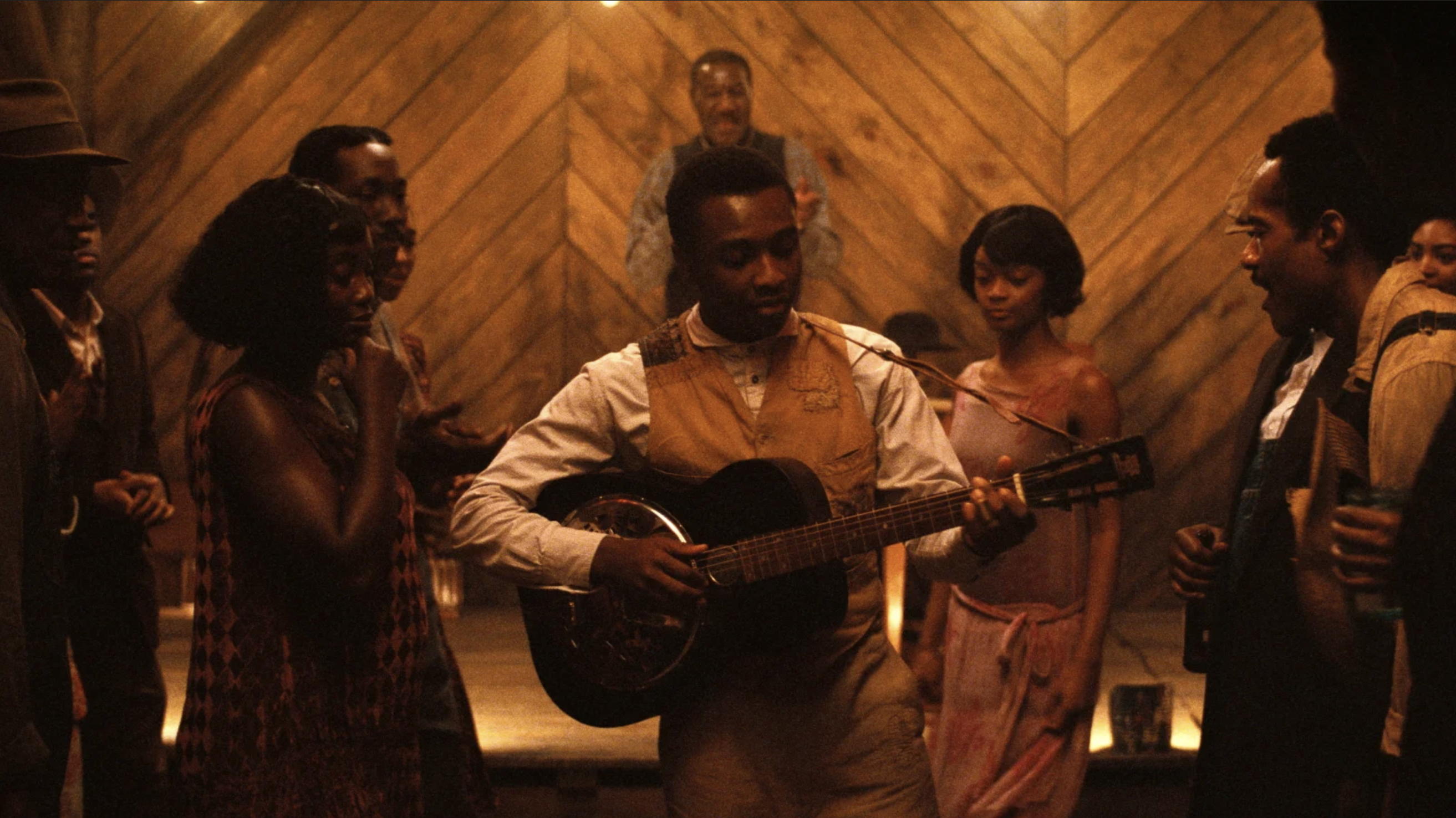 A young Black man plays a guitar while other people listen around him in a barn dance setting.