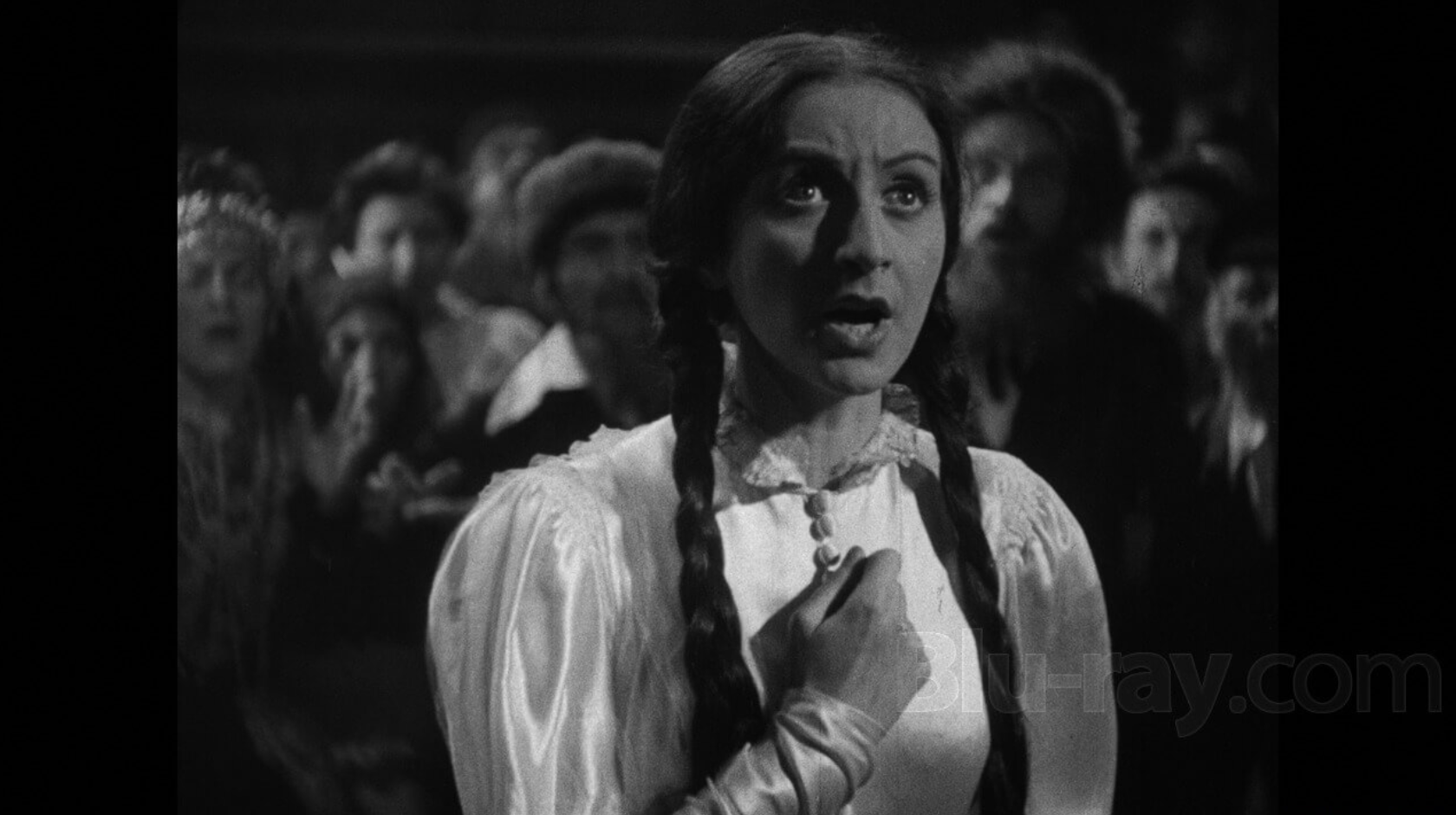 Young woman, in white dress with two long braids, in the midst of a crowd speaking with hand on heart.
