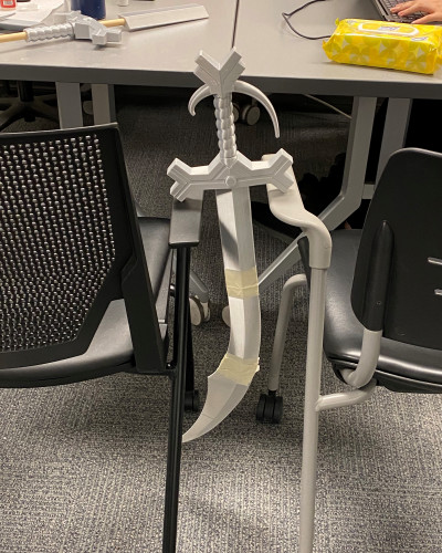 Silver 3D printed simitar sword hanging by the hilt between two chairs to allow the glue between the joints of each piece to dry efficiently
