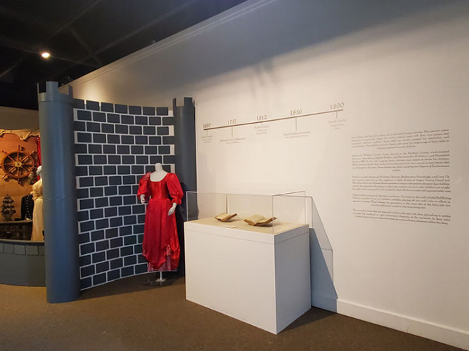 This display shows a bright red princess dress on a mannequin in front of a black, faux brick wall. Two of the SCRC fairytale books are displayed open in a case, with a timeline from 1607 to 1890 on the wall behind.