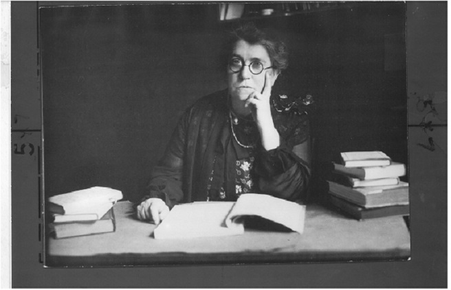 Black and white photograph of Emma Goldman sitting at a desk surrounded by books