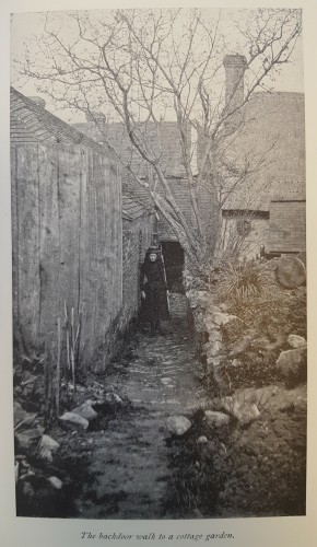 Black and white photograph of a girl standing in a narrow path to a cottage door.