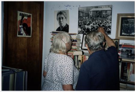 Photo of two people with their backs to the camera, one pointing at a photo of Emma Goldman that is hanging on the wall