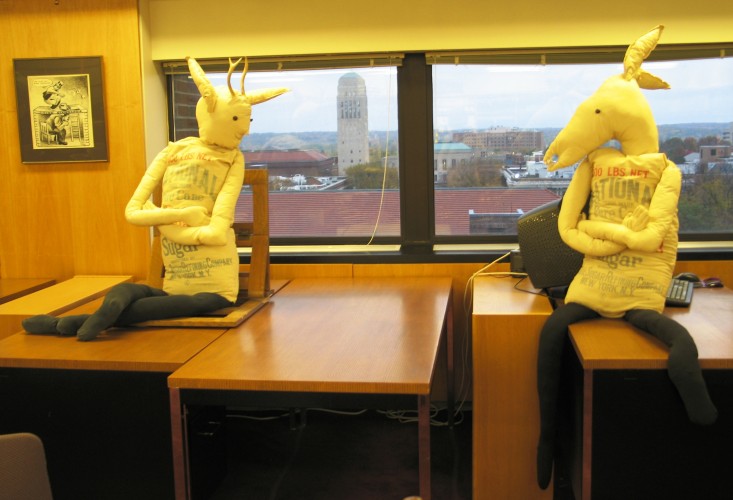 Two nearly-human sized figurines made of floursacks with animal heads, sit on top of tables in front of a window