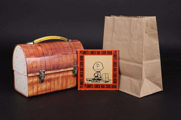 brown lunch box shaped and designed like a loaf of bread standing beside square cookbook with cartoon on cover and paper lunch sack
