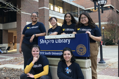 A group of library student employees wearing U-M Library t-shirts, smiling, and standing around the Shapiro Library sign.