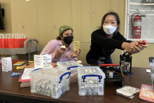 Two students, wearing masks, holding up a cupcake and making pinback buttons.